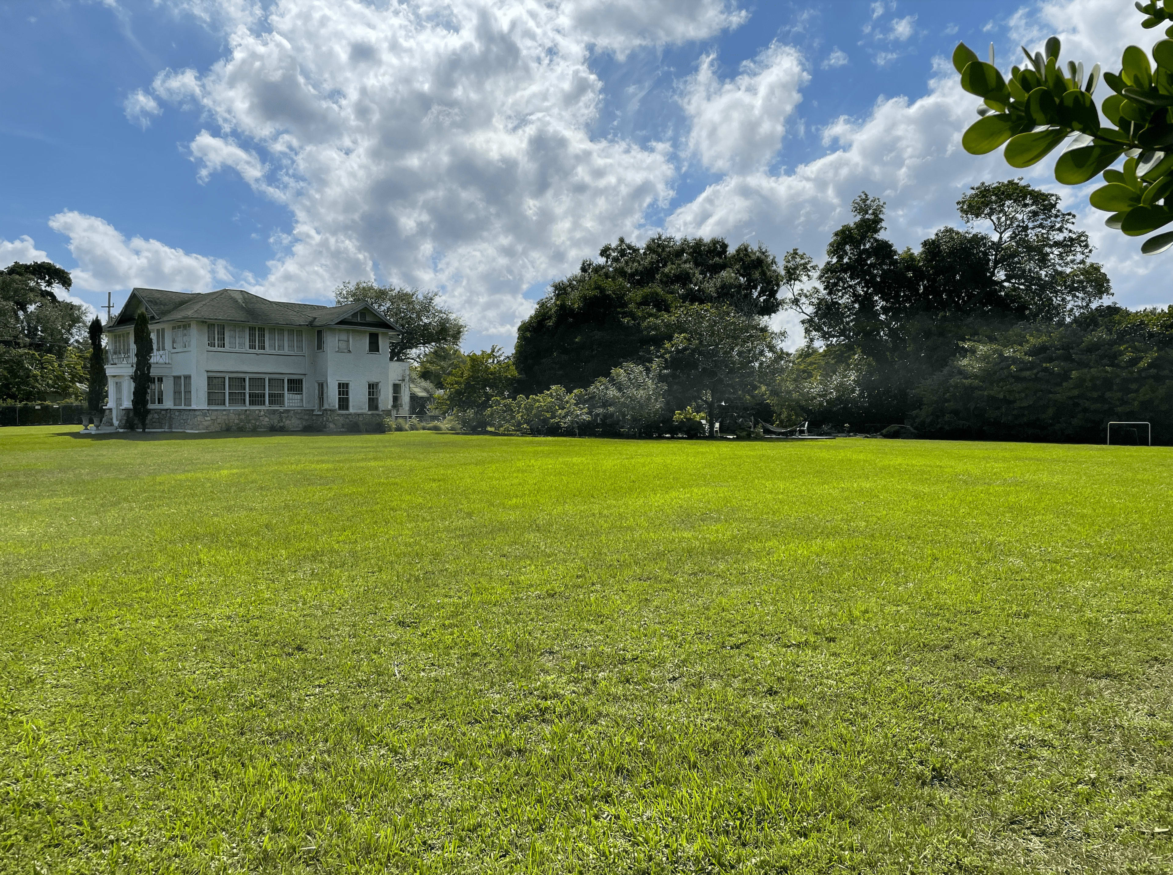 A large, two-story white house is situated on a grassy field surrounded by trees under a partly cloudy sky.