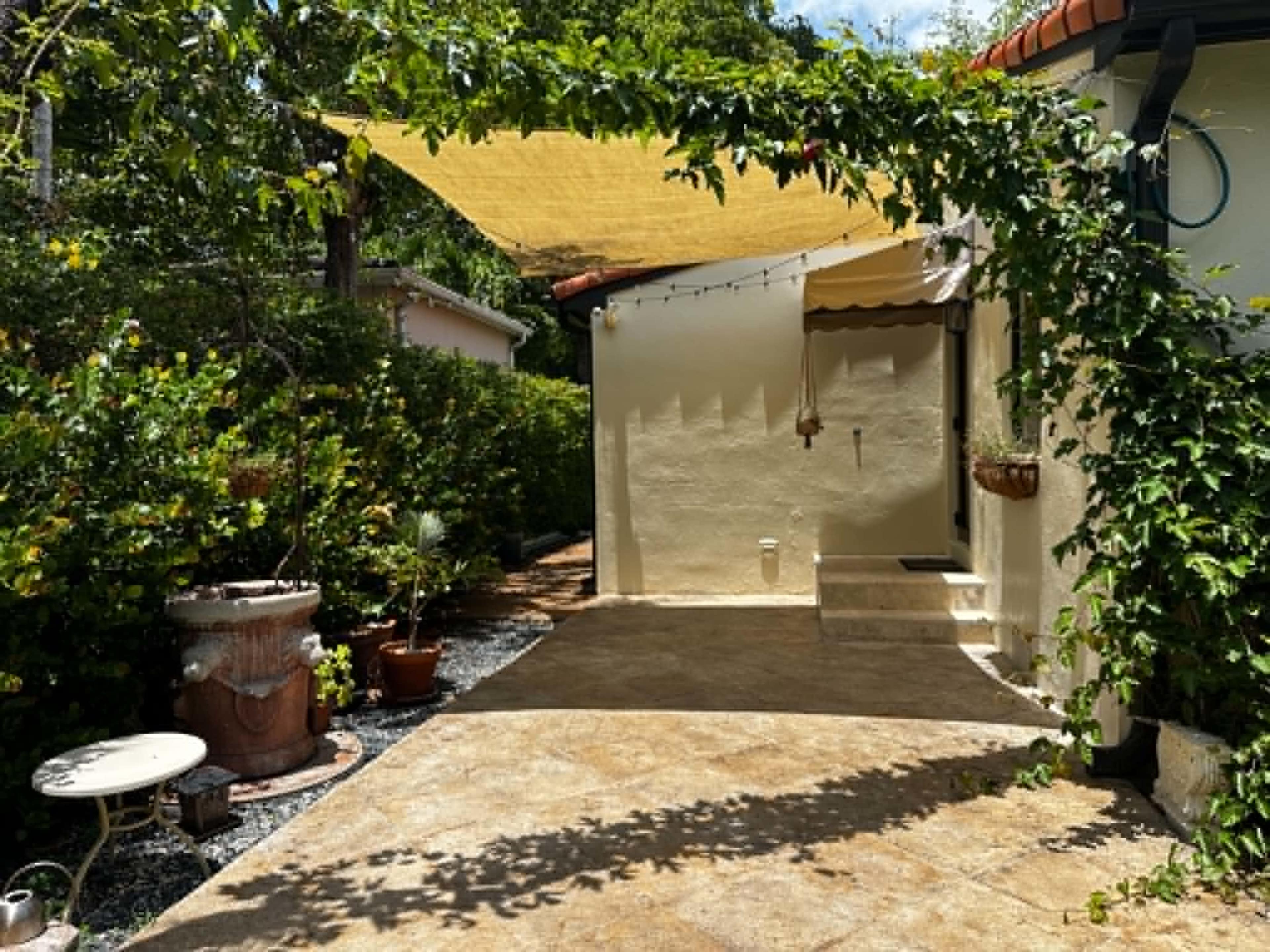 The image shows a landscaped outdoor area featuring a terracotta tiled floor, green foliage, and a yellow canopy overhead.