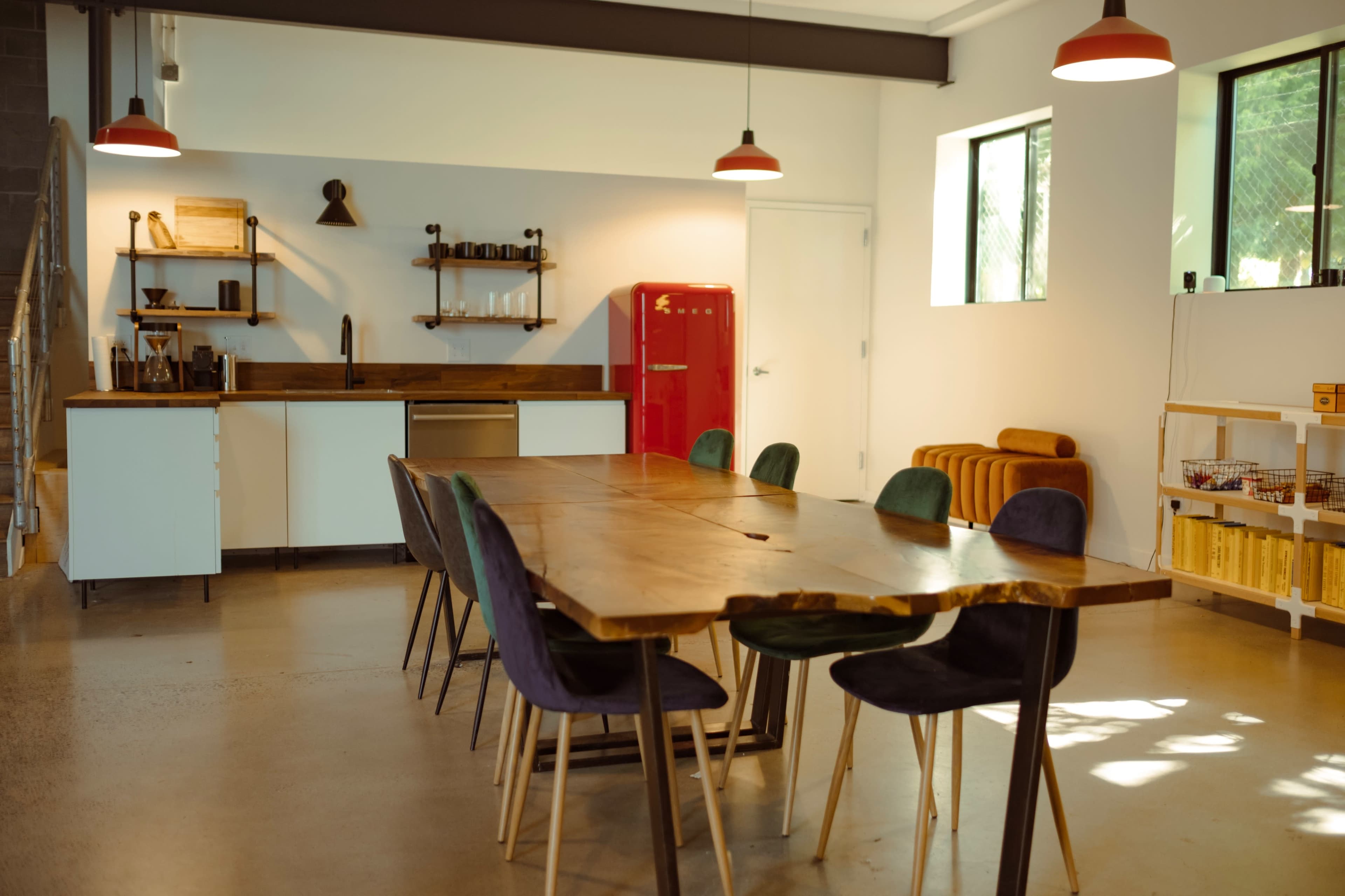 A spacious dining area features a long wooden table surrounded by colorful chairs, with a modern kitchen in the background and a red refrigerator.