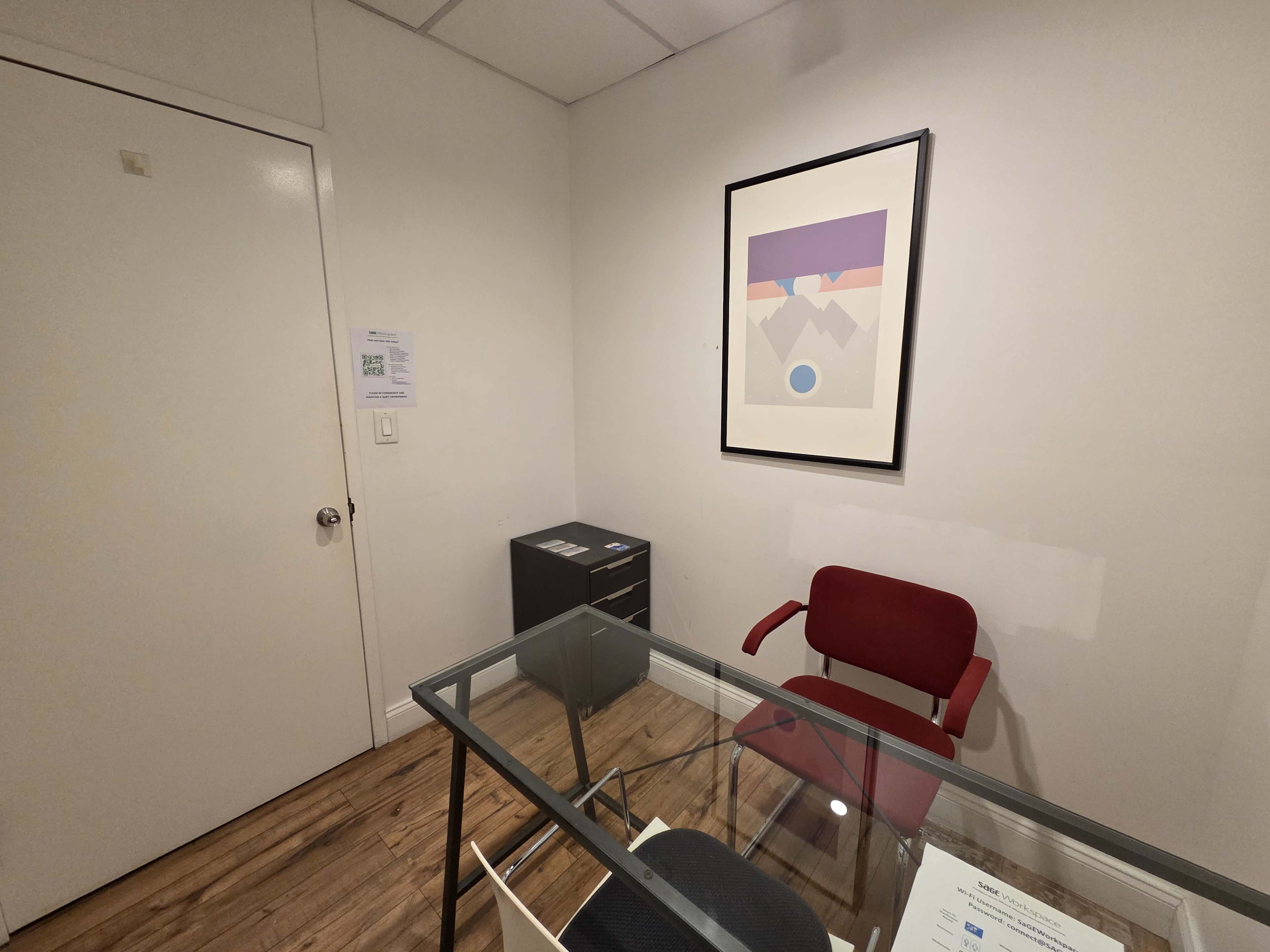 The image shows a small, minimally furnished office with a glass table, a red chair, and a framed artwork on the wall.