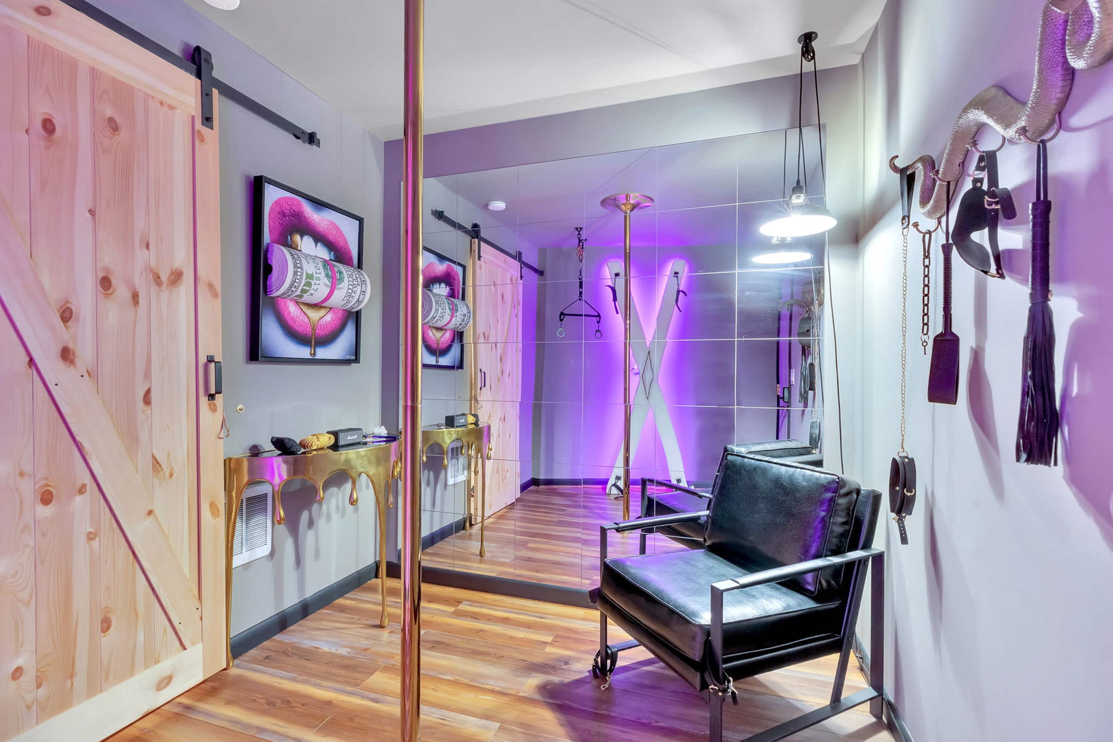 A modern room with a mirrored wall, a black chair, a pole, and purple lighting, featuring a wooden door and framed artwork on the walls.