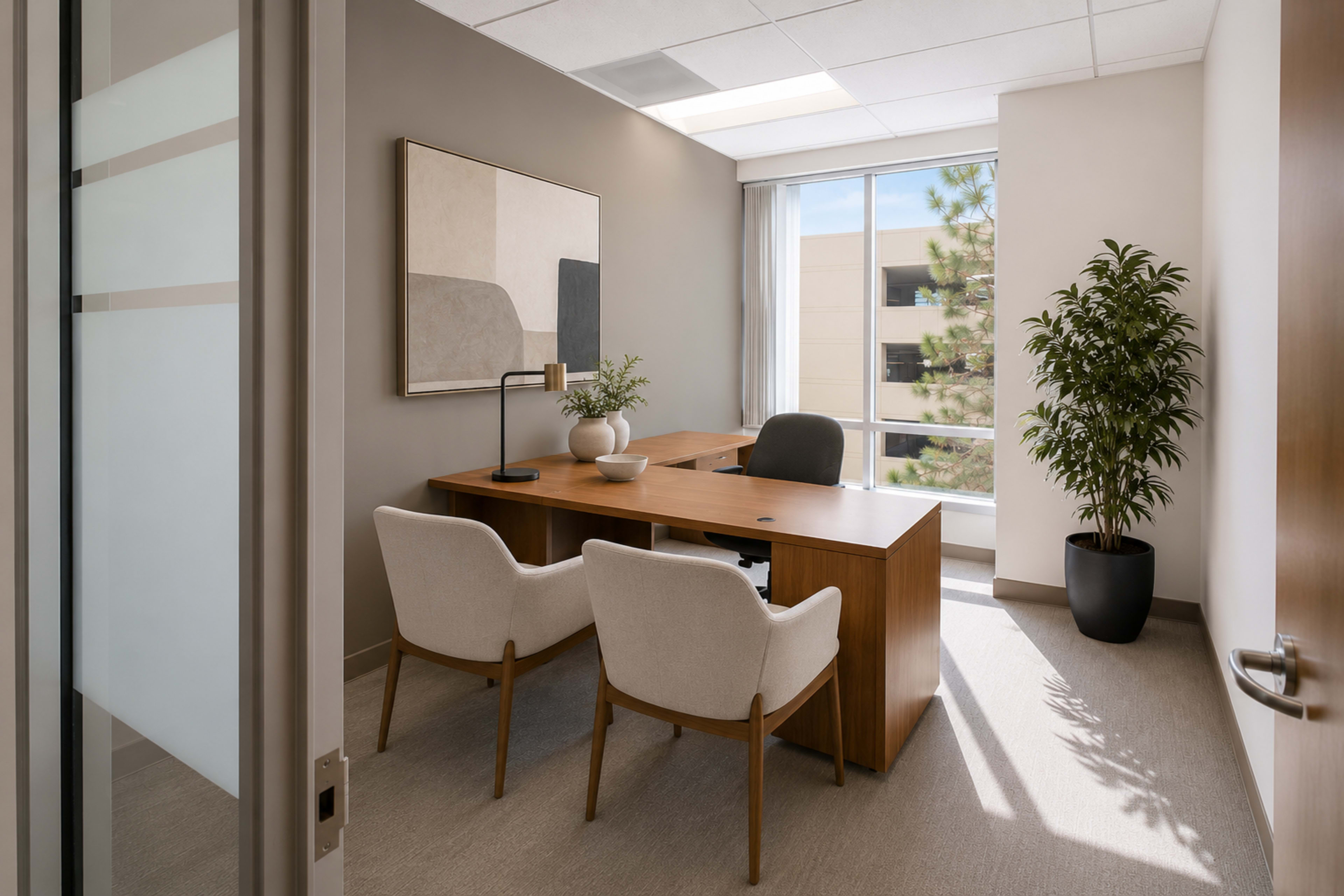 A modern office space features a wooden desk, two upholstered chairs, a decorative plant, and a large window allowing natural light.