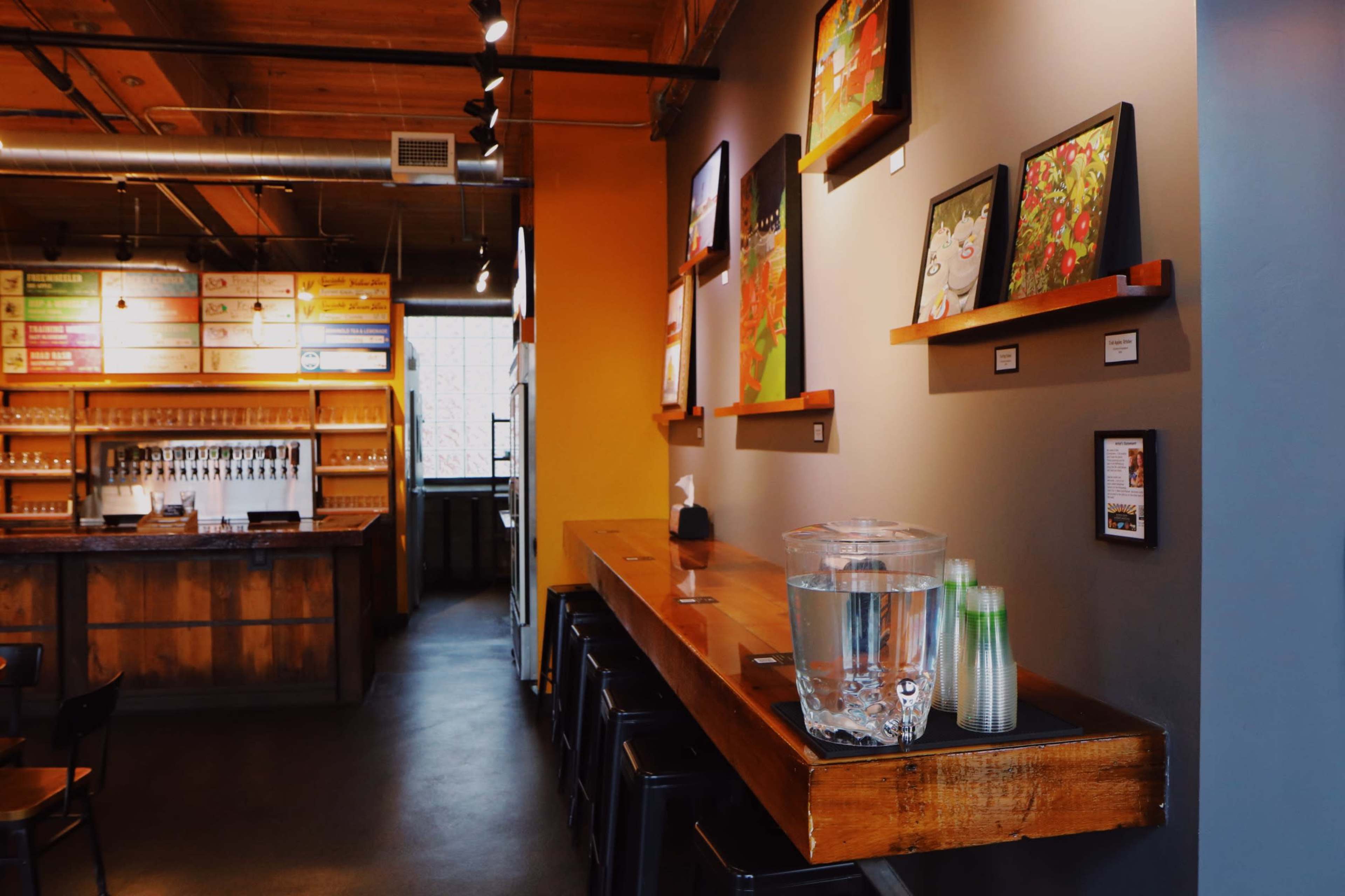The interior of a modern café featuring wooden shelves with colorful artwork, a long wooden counter, and a beverage station with glass pitchers and cups.