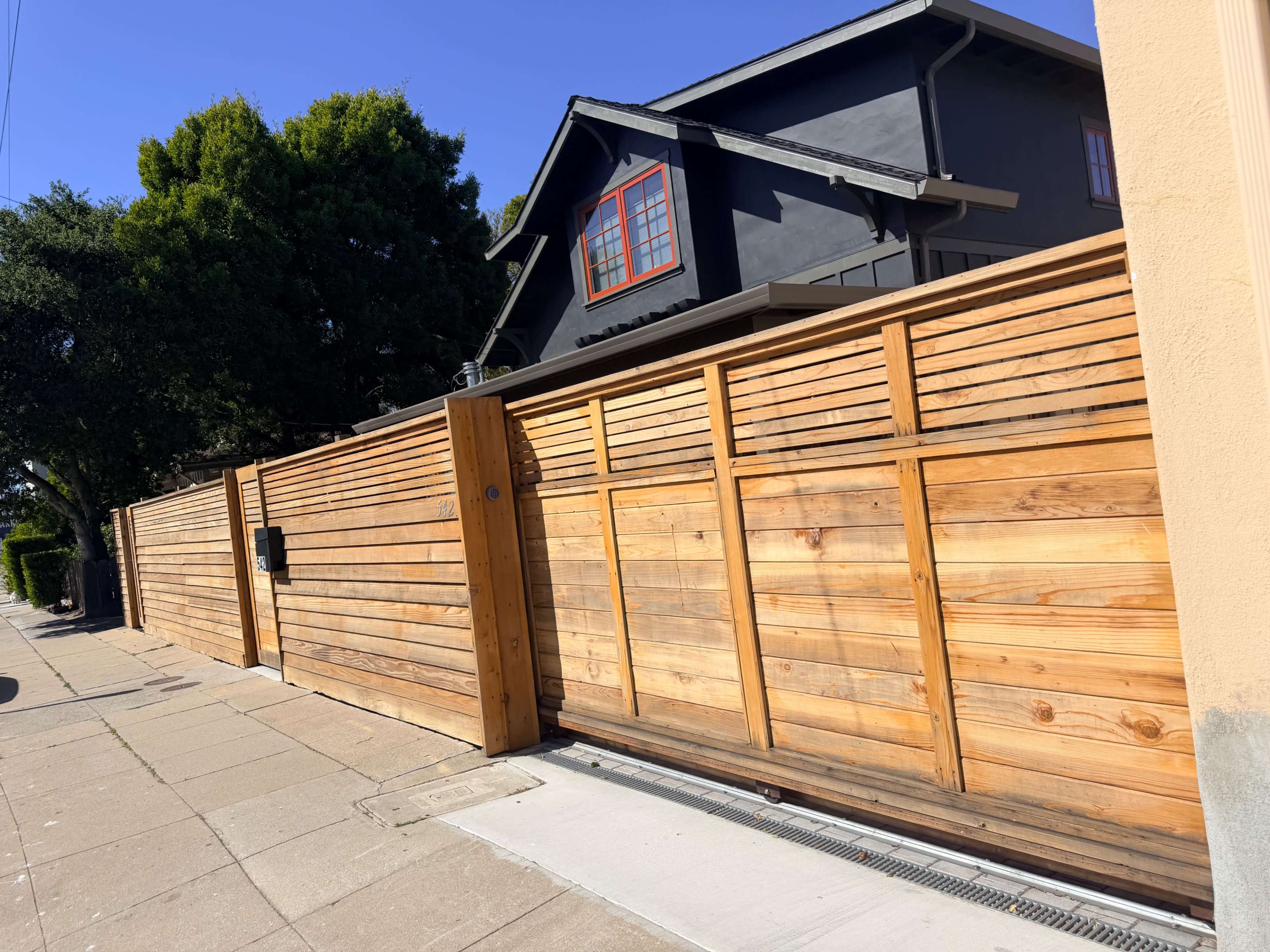 A wooden fence encloses a modern house with a dark exterior and red window trim under a clear blue sky.