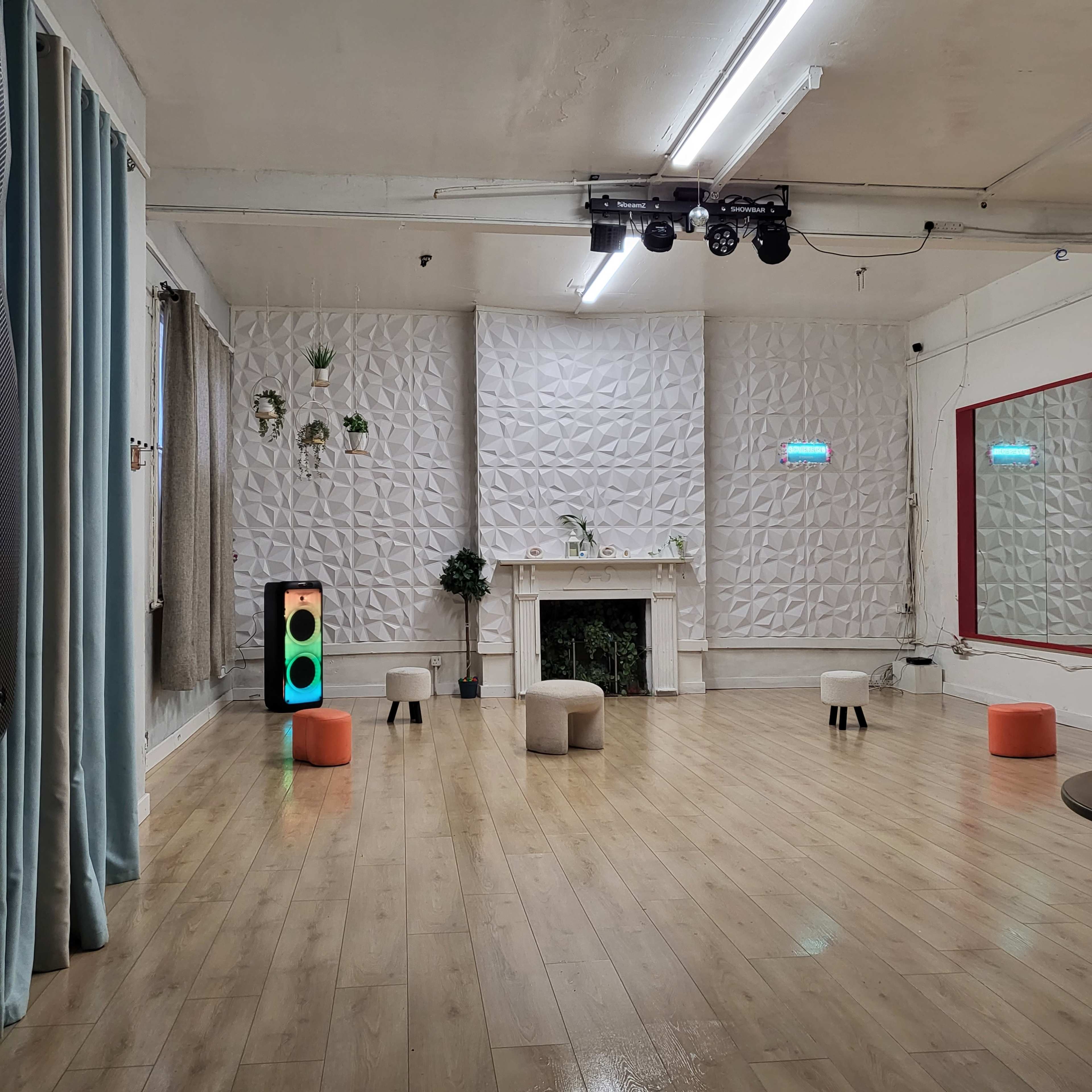 The image shows a spacious, well-lit room with wooden flooring, featuring a decorative wall, a fireplace, several small stools, and colorful lights.