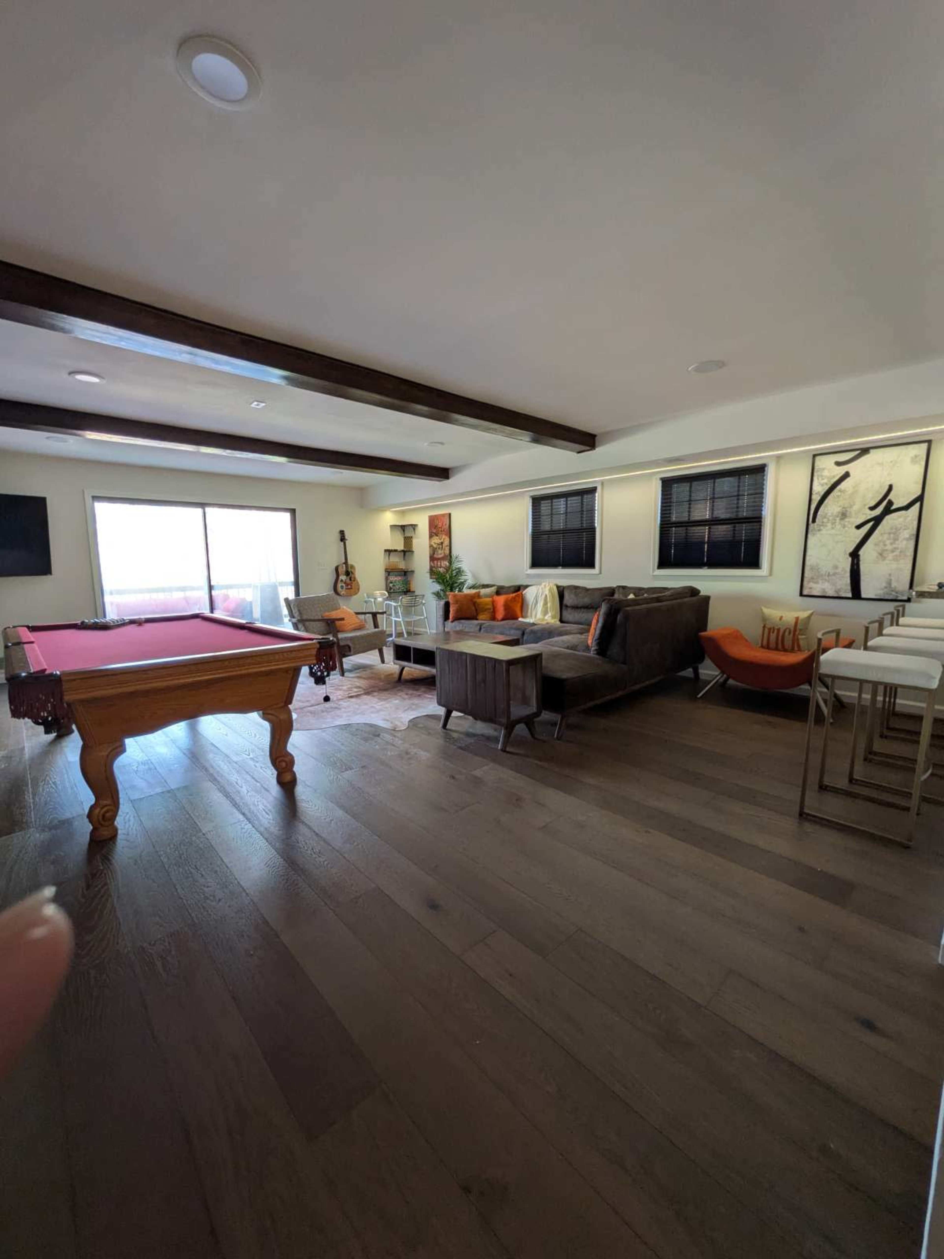 The image shows a living area with a pool table, a seating area featuring a sofa and chairs, and large windows providing natural light.