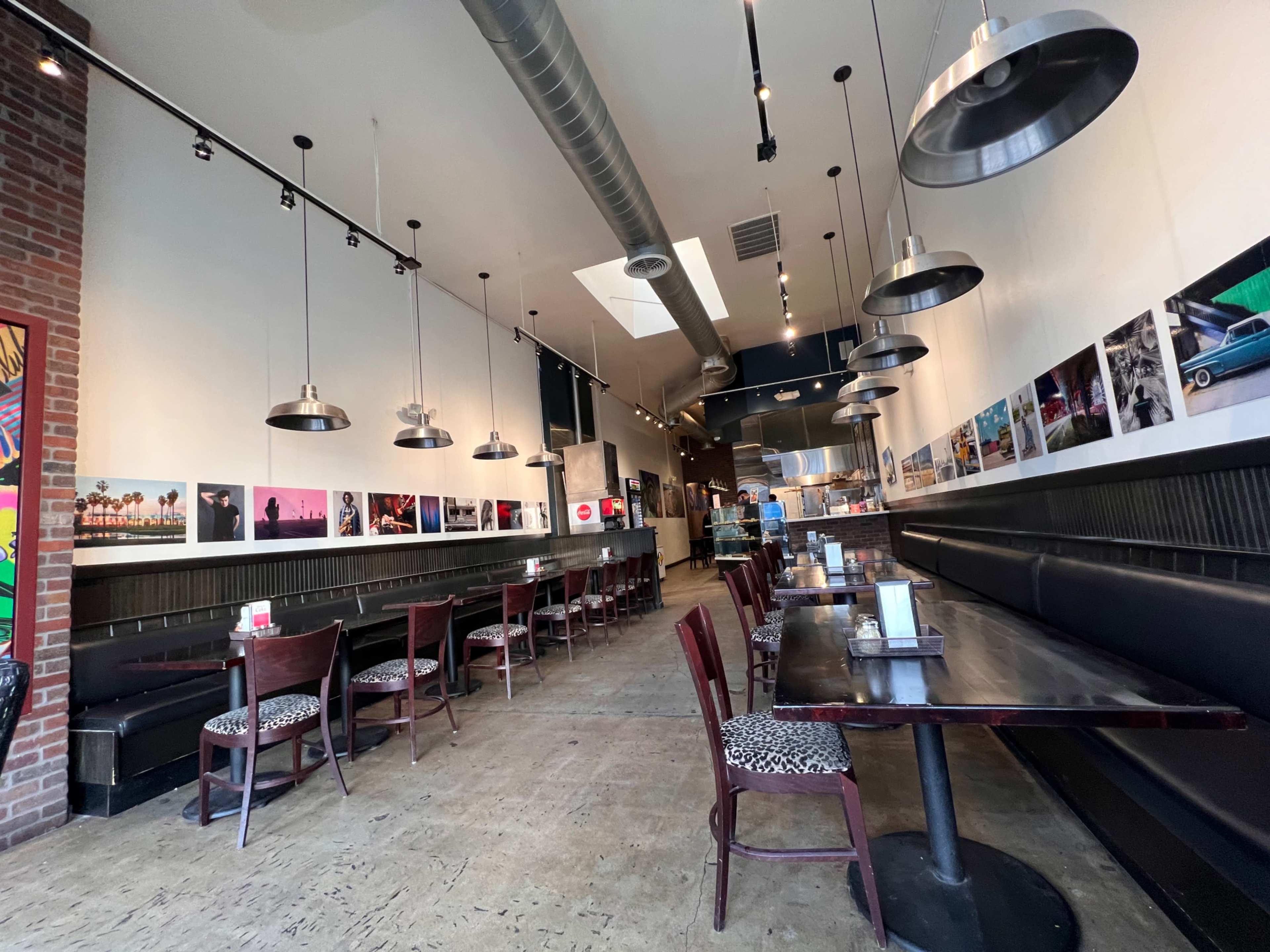 The image shows a narrow restaurant interior with long tables, patterned seating, and pendant lights, featuring framed photographs on the walls.