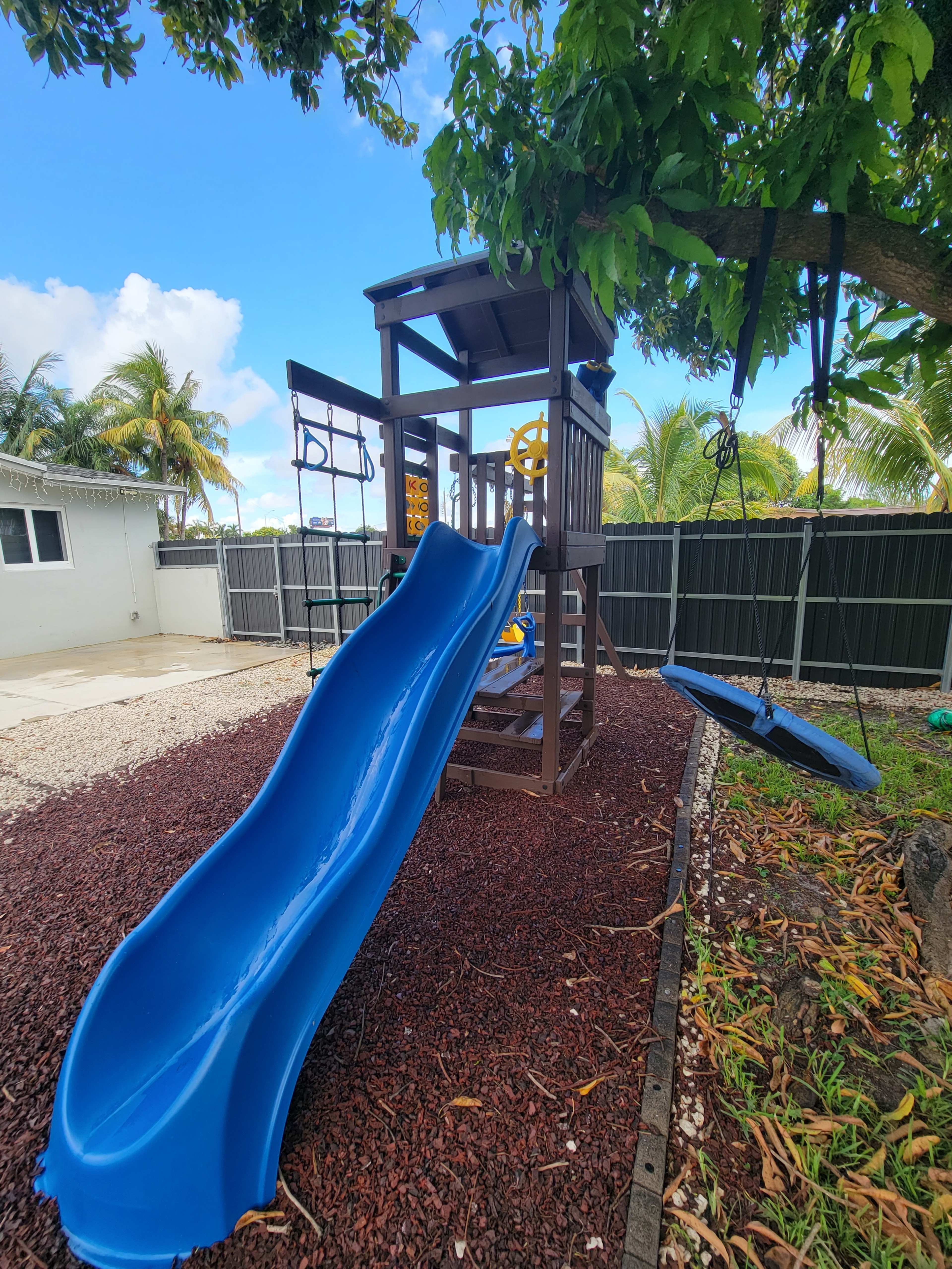 The image shows a playground structure with a blue slide, swings, and a climbing frame set on gravel in a backyard.