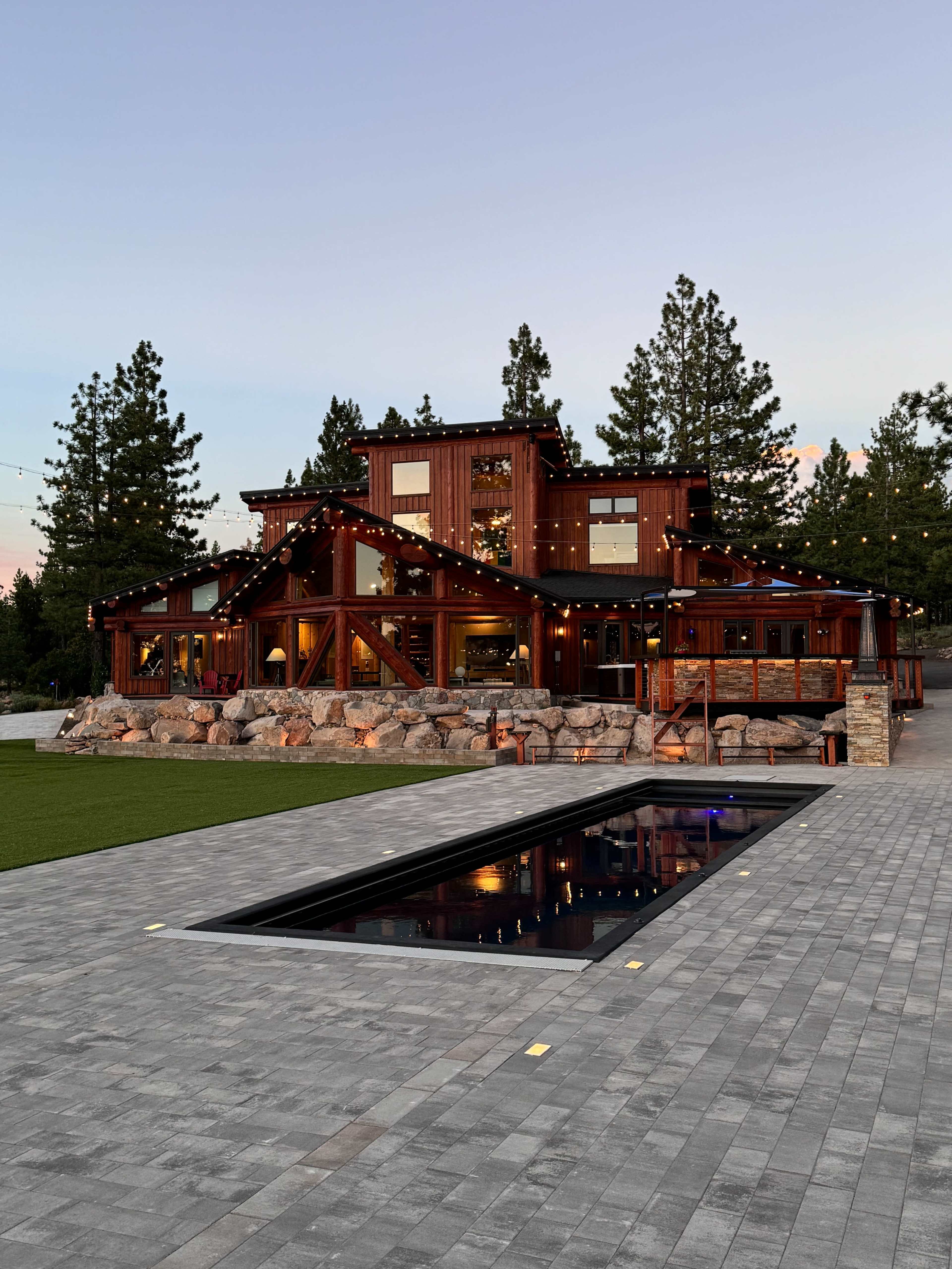 A modern wooden house with large windows and a stone exterior is surrounded by landscaped grounds and features a long, rectangular pool in the foreground.