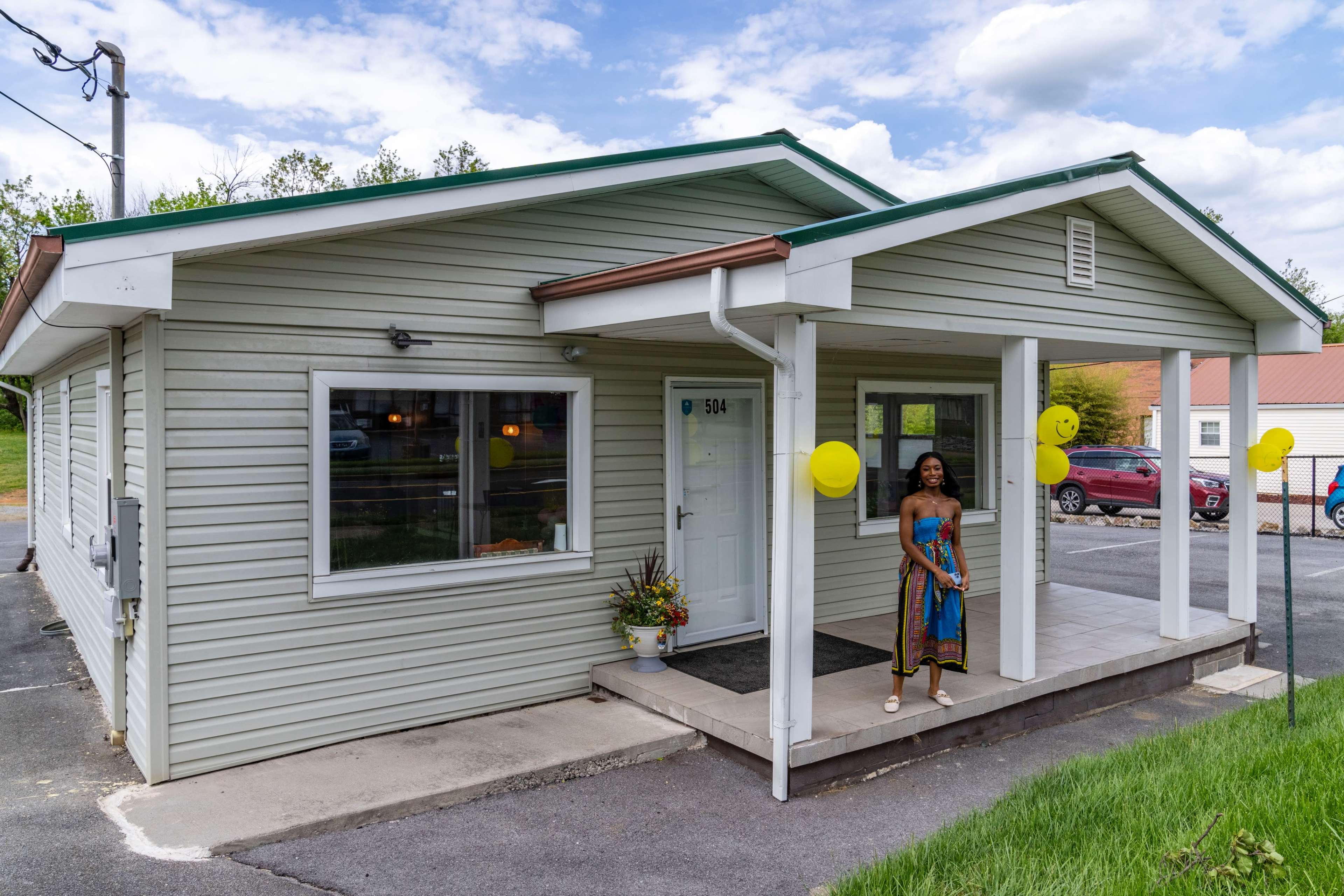A woman stands outside a single-story green building with a front porch, yellow balloons, and a neatly maintained landscape.