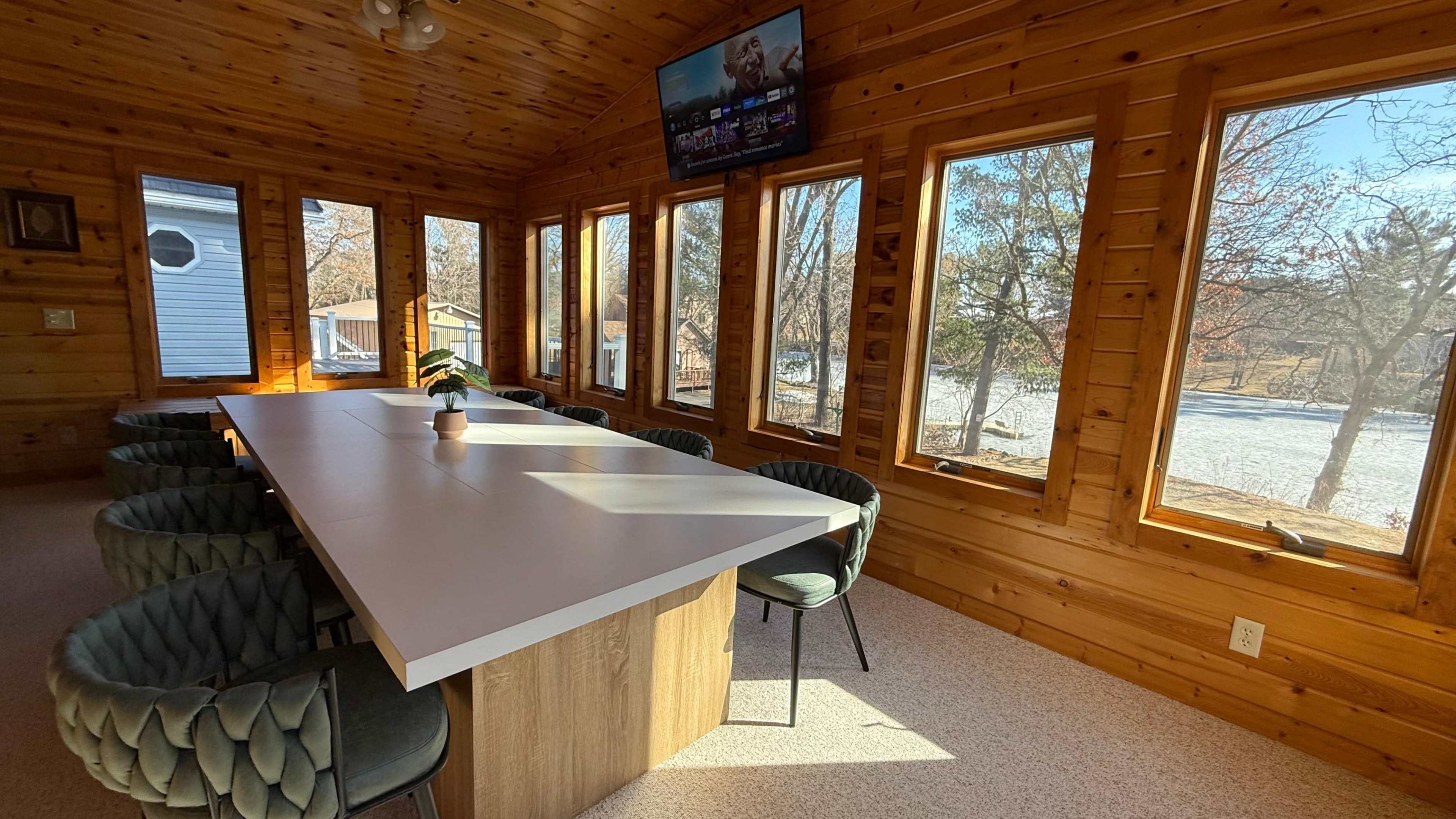A spacious sunroom features a large white table surrounded by six upholstered chairs, with large windows providing views of the outdoors.