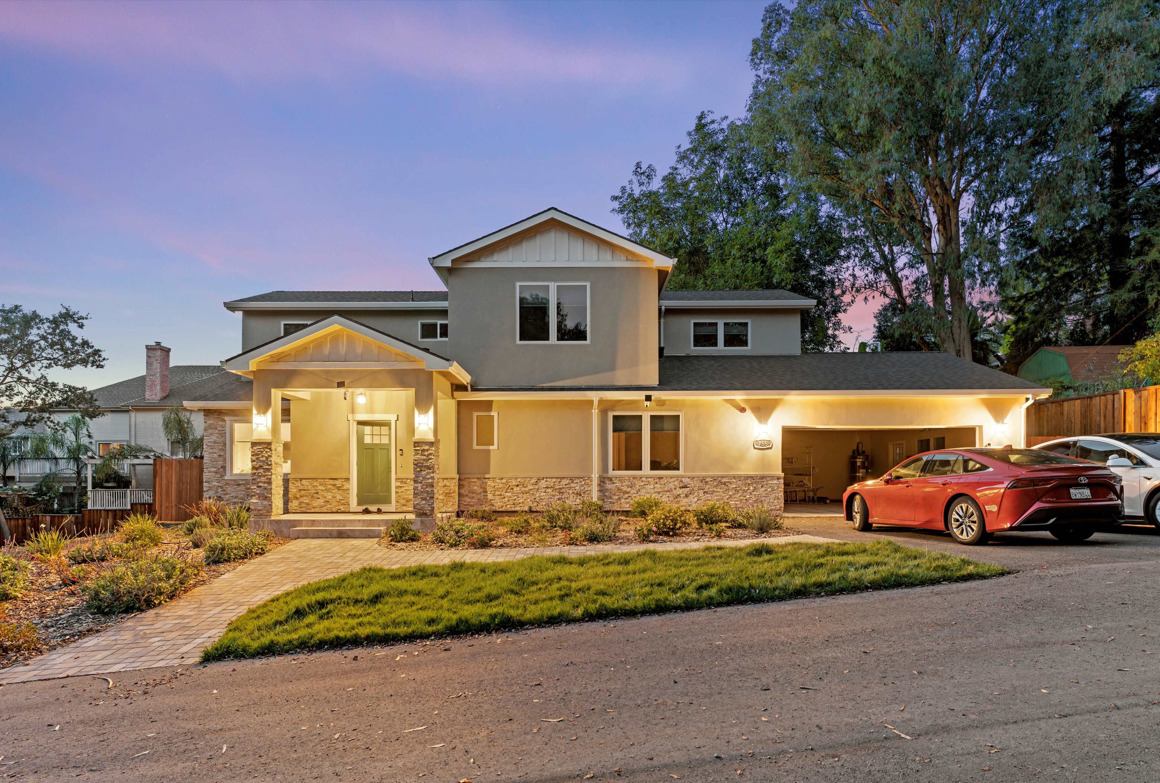 A two-story house with a landscaped front yard and a driveway showcases a red car parked beside it during twilight.