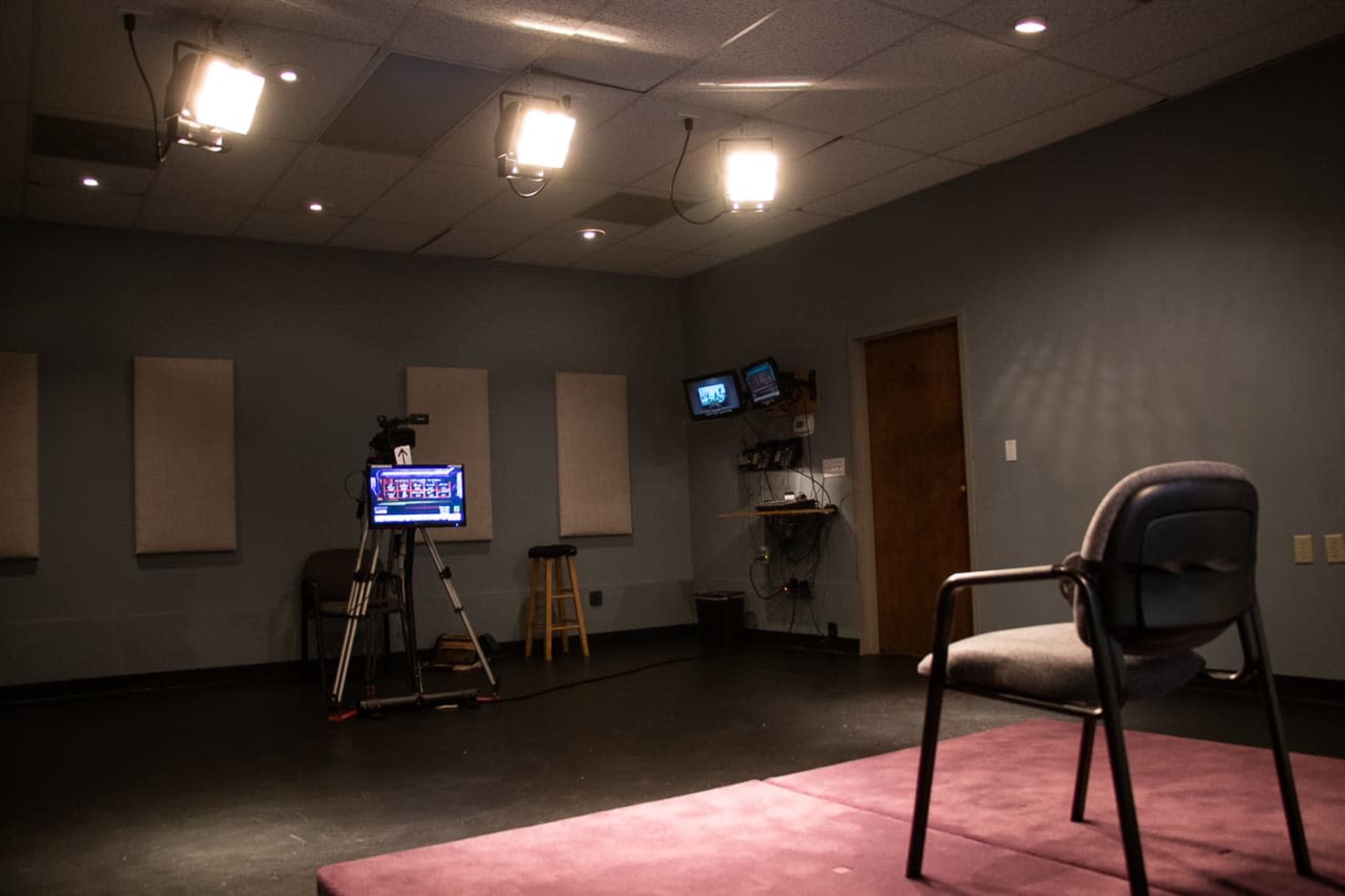 A television studio setup with a camera on a tripod, a monitor displaying graphics, and an empty chair placed on a purple mat.