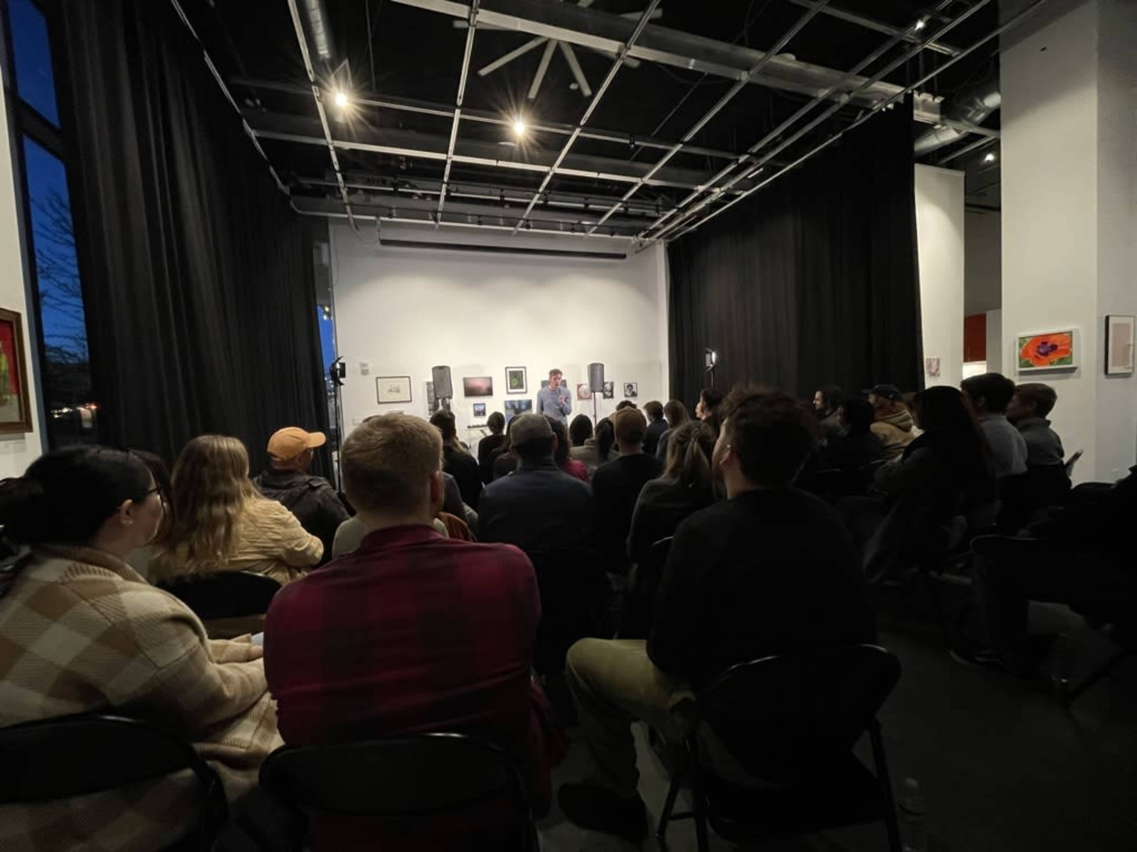 A large group of people sits in a dimly lit gallery, facing a speaker at the front of the room.