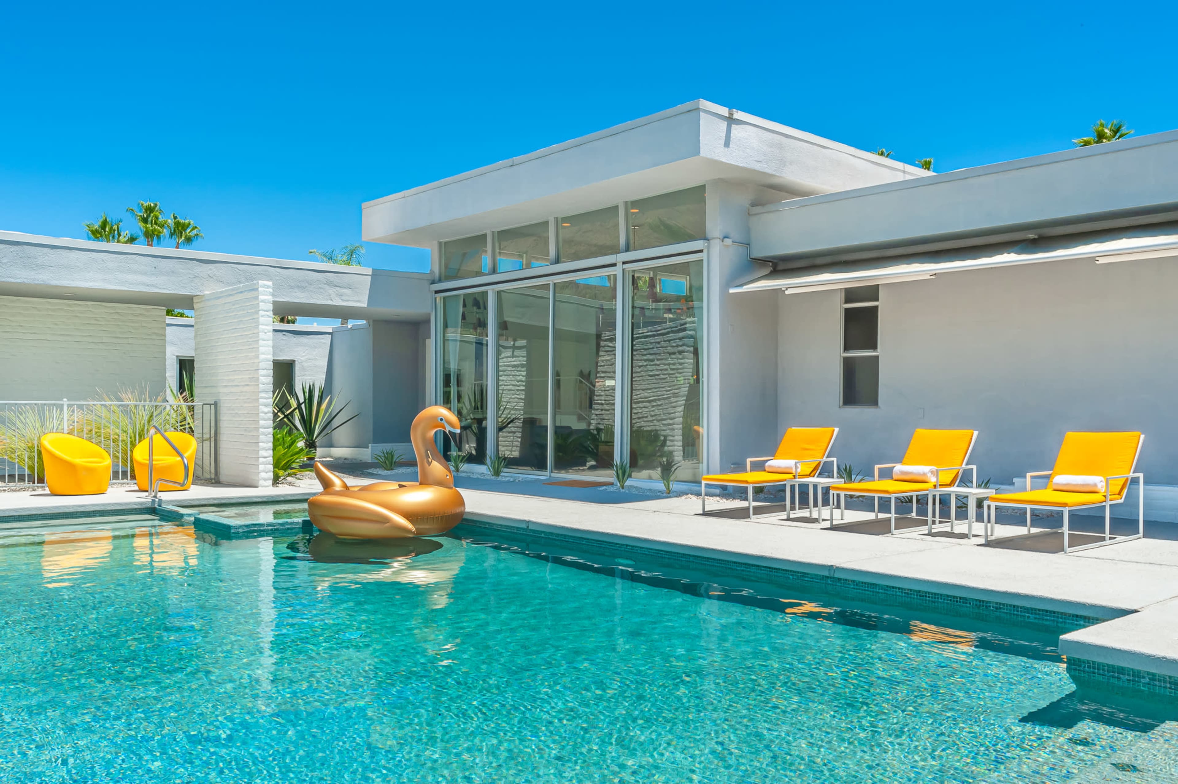 A modern pool area features bright orange lounge chairs, a large inflatable swan, and glass walls that allow for views into the interior spaces.