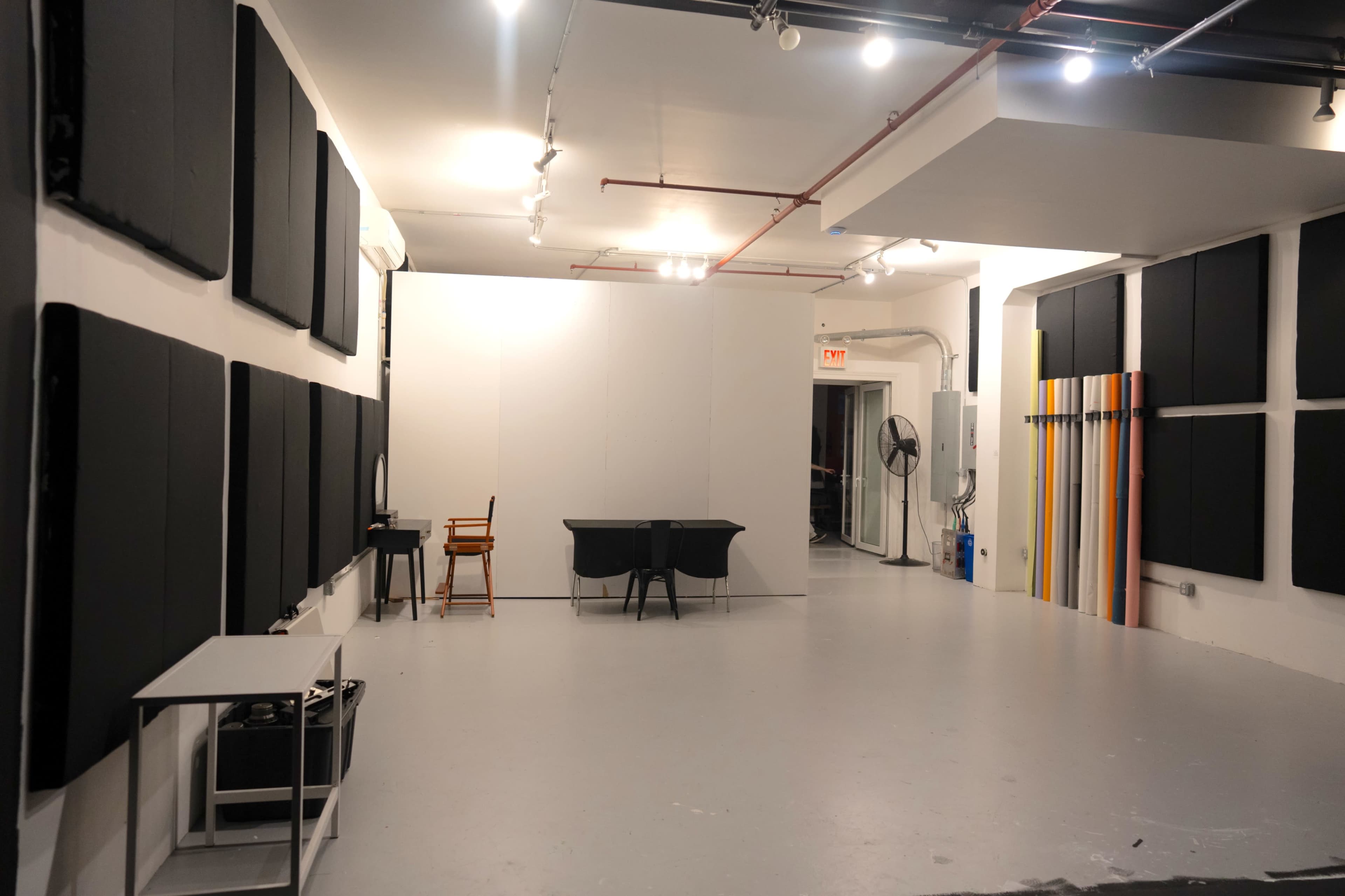 The image shows an interior space with soundproofing panels on the walls, a table with chairs at the center, and various colorful boards leaning against one wall.