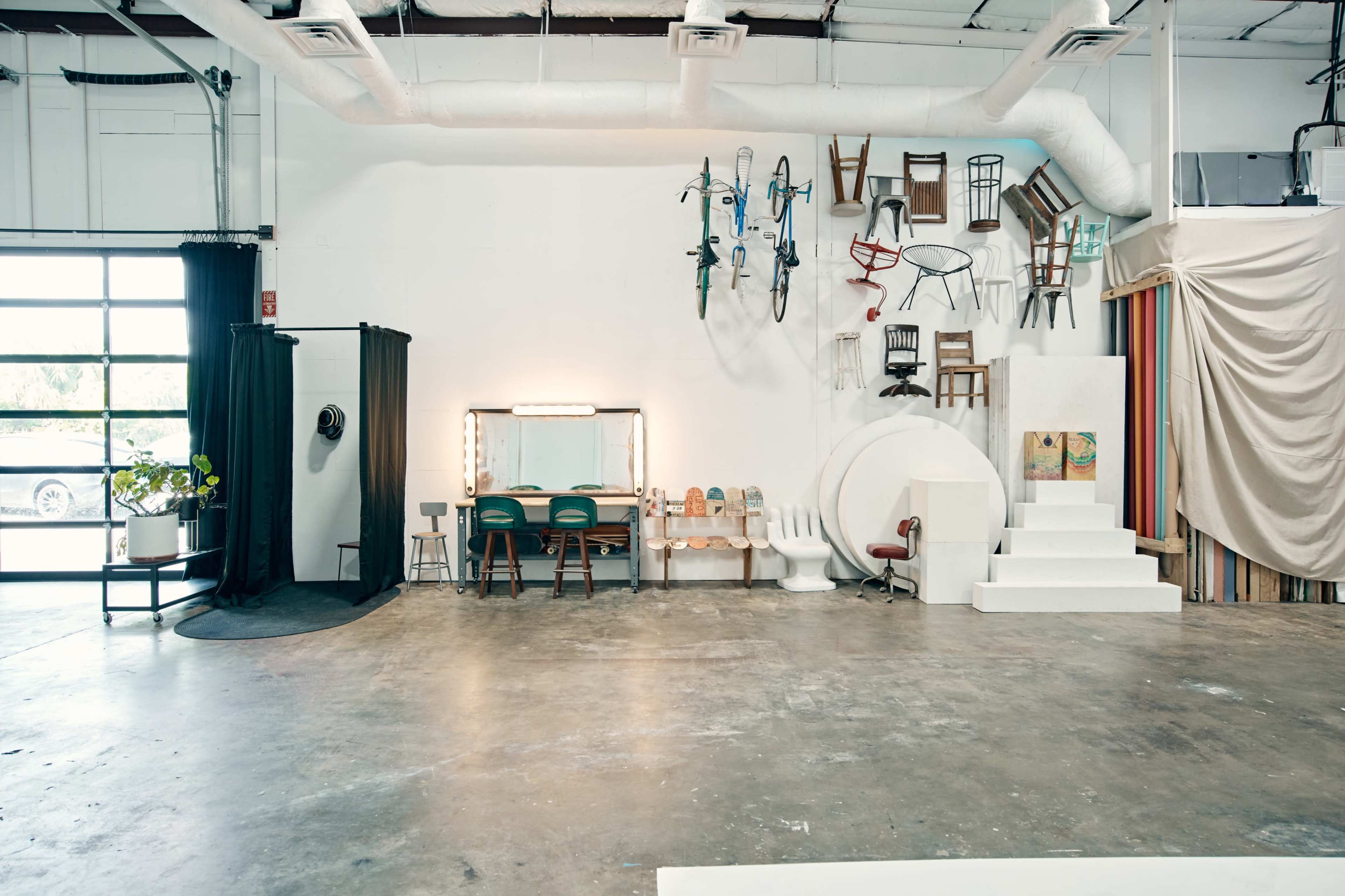 A spacious studio with various chairs and bicycles mounted on a white wall, a dressing table with two chairs, and a series of white pedestals on a polished concrete floor.