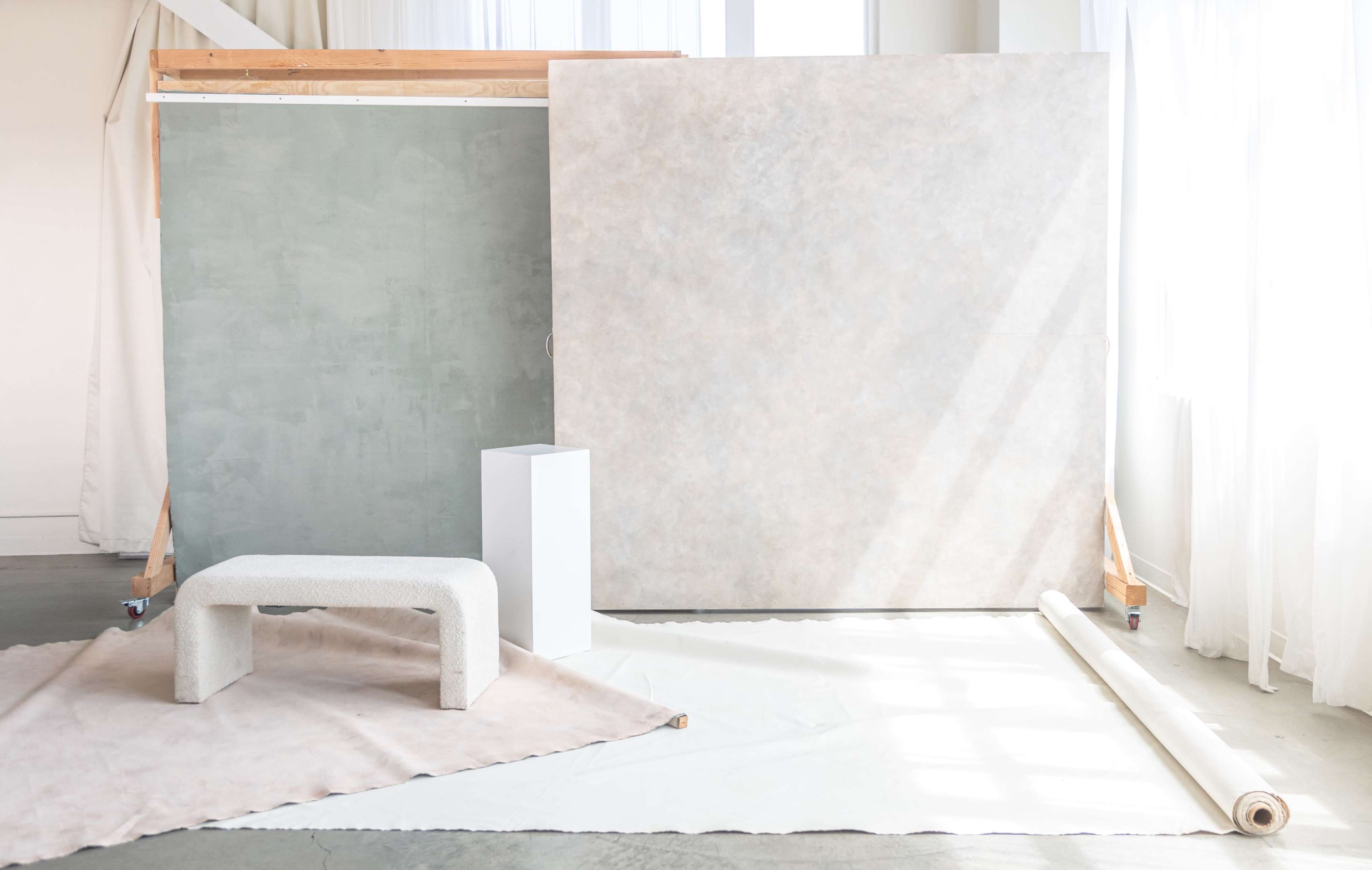 The image features a minimalist studio setup with two large textured backdrops, a sculptural bench, and a rolled-up fabric on the floor.