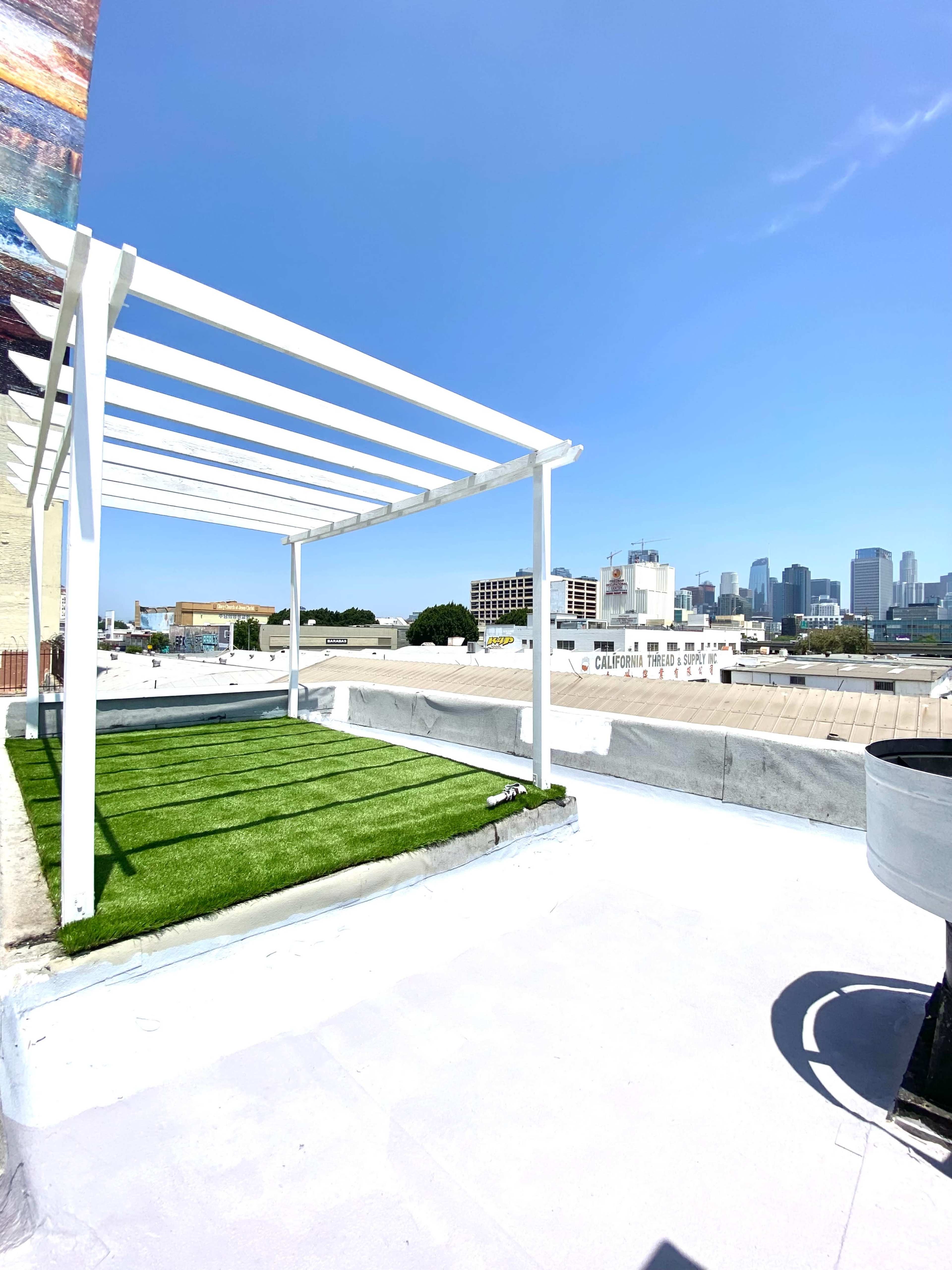 A white pergola stands over a green turf area on a rooftop with a city skyline in the background.