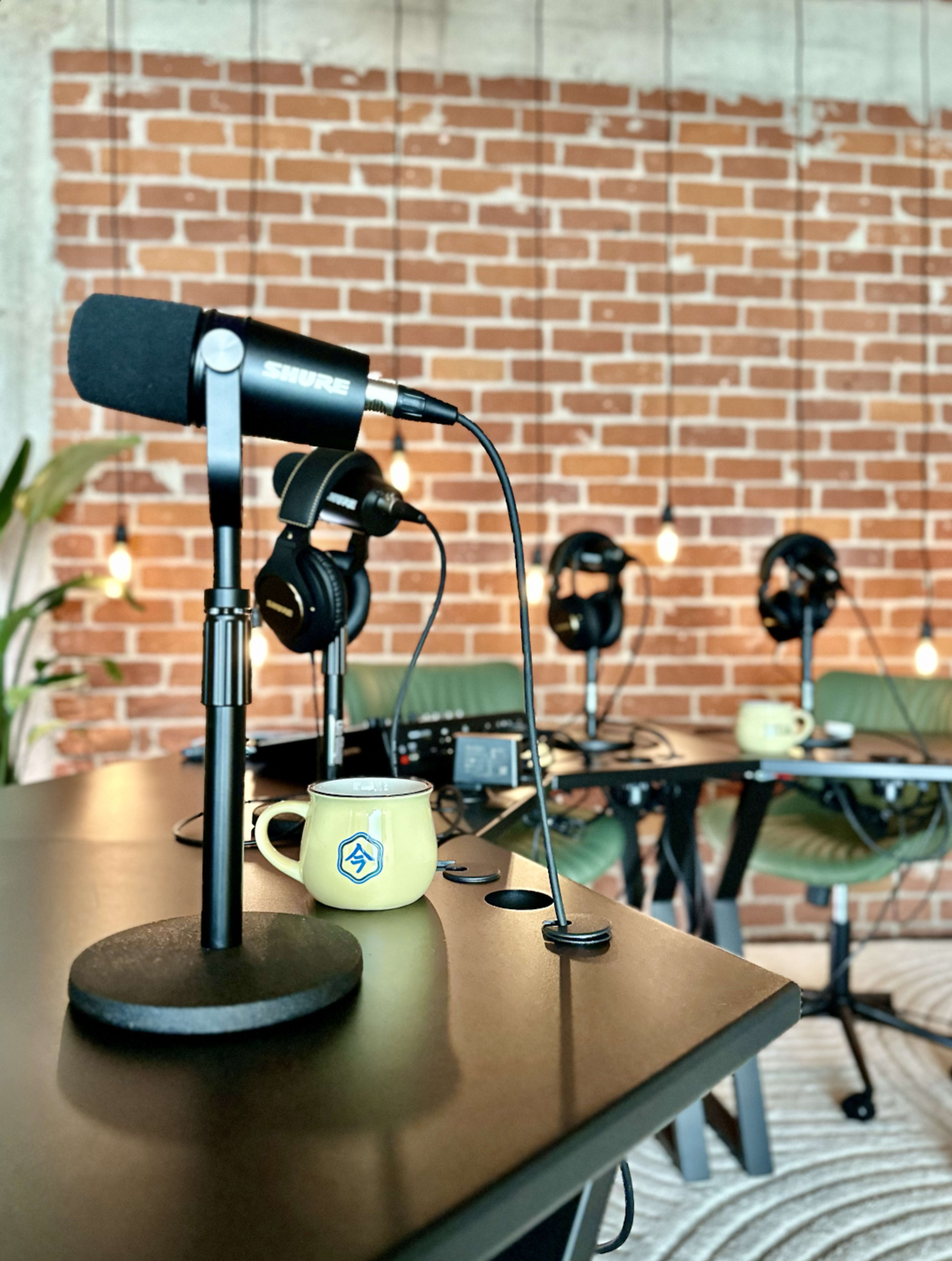 A microphone and a coffee mug sit on a desk in a room with exposed brick walls and headphones hanging nearby.