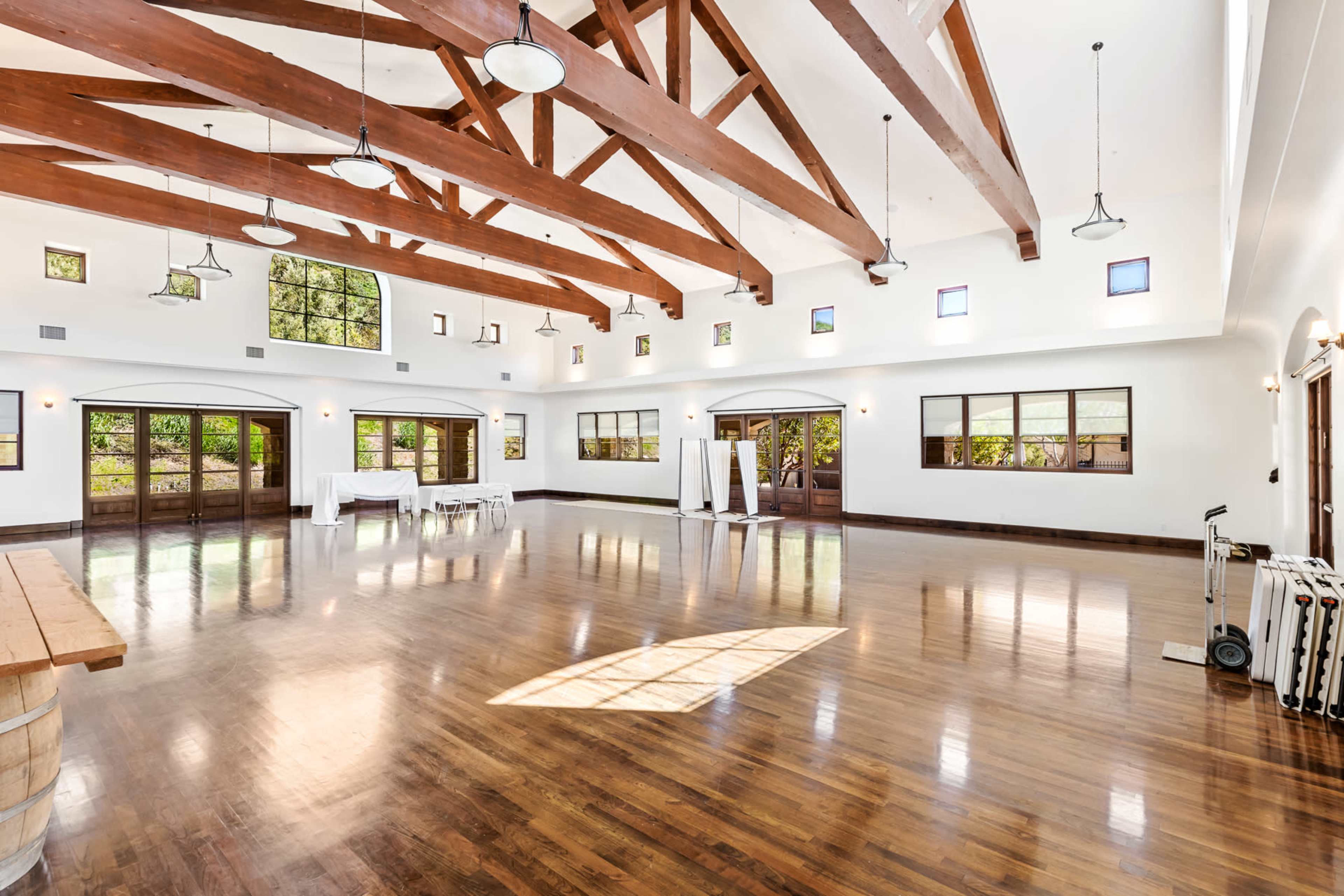 The image shows a spacious event hall with high wooden beams, large windows, and polished wooden floors.