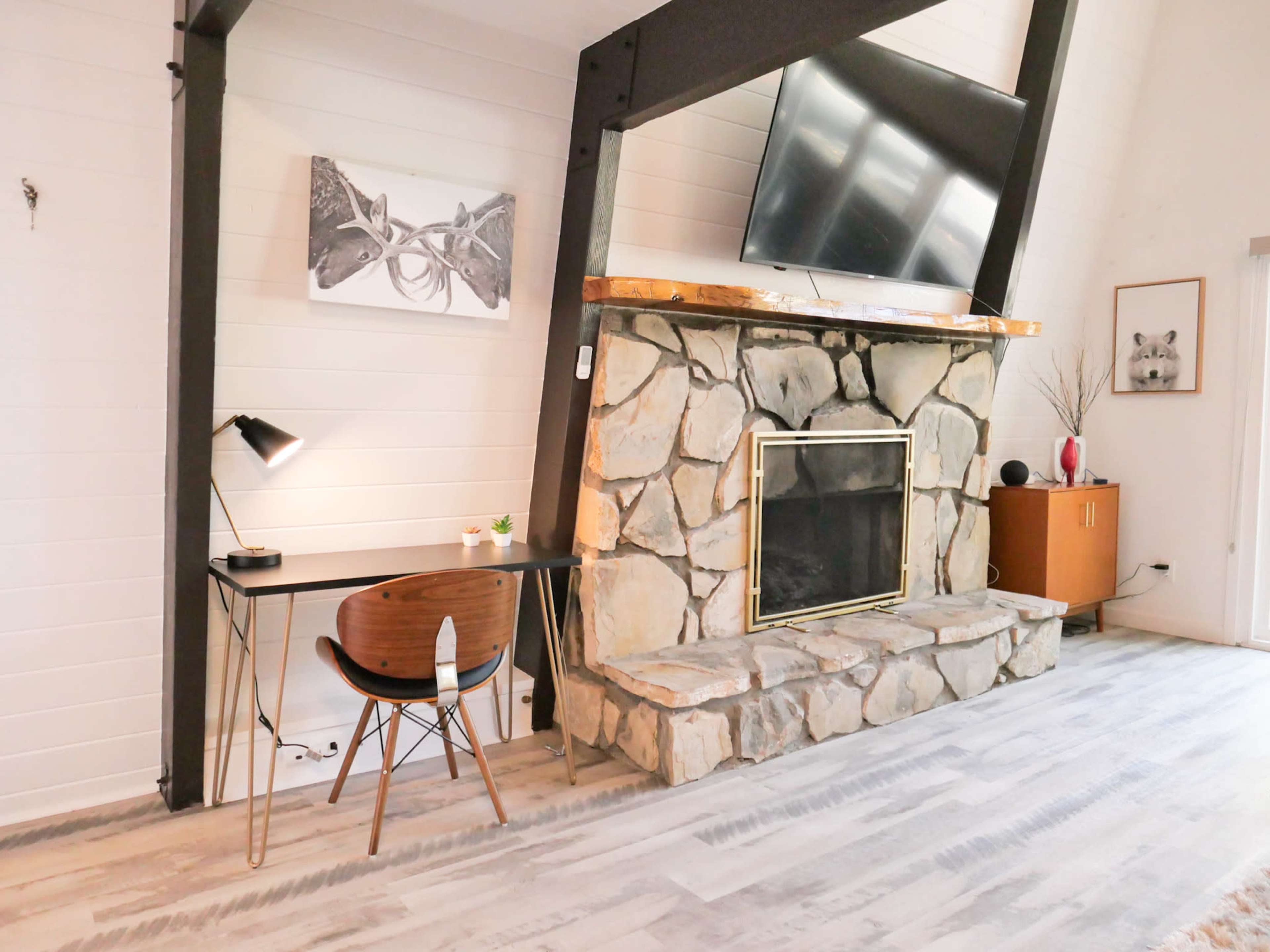 The image shows a room with a stone fireplace, a wooden desk with a chair, and various artworks on the walls.