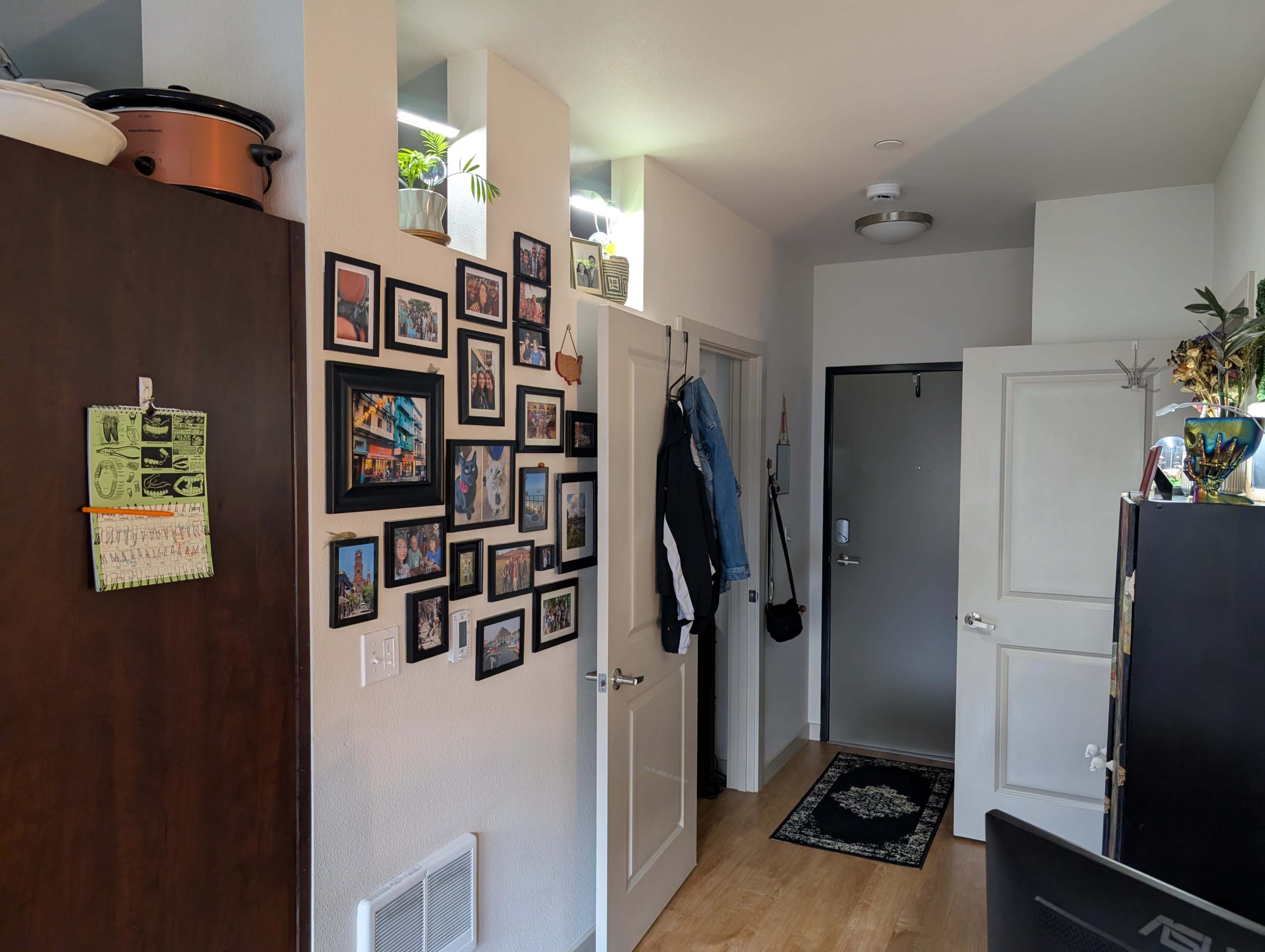 The image shows the interior of a small living space with a wall decorated with framed photos, a coat hanging on a door, and a dark cabinet in the corner.