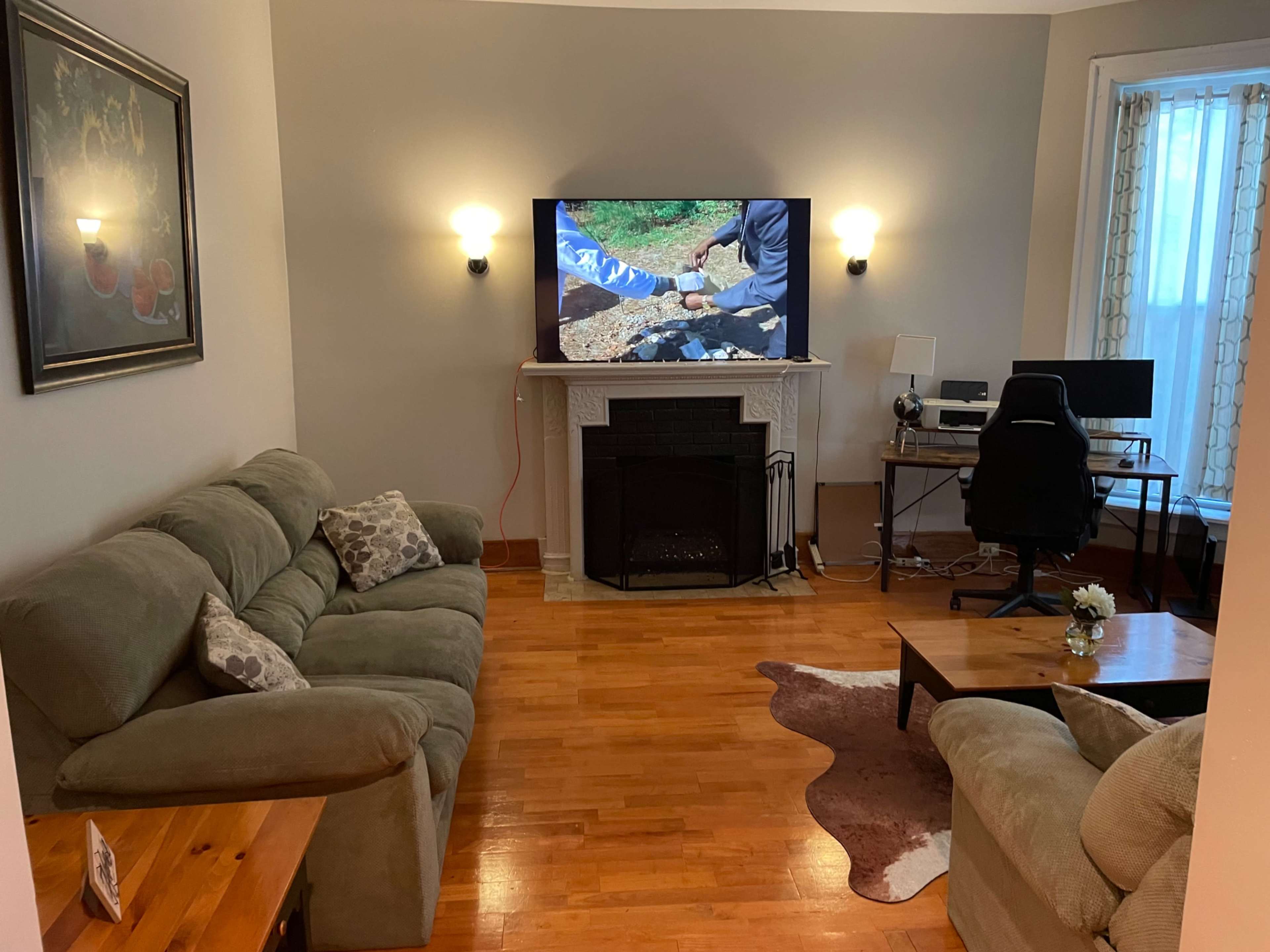 A living room features a couch, armchair, wall-mounted TV above a fireplace, and a desk with a chair.