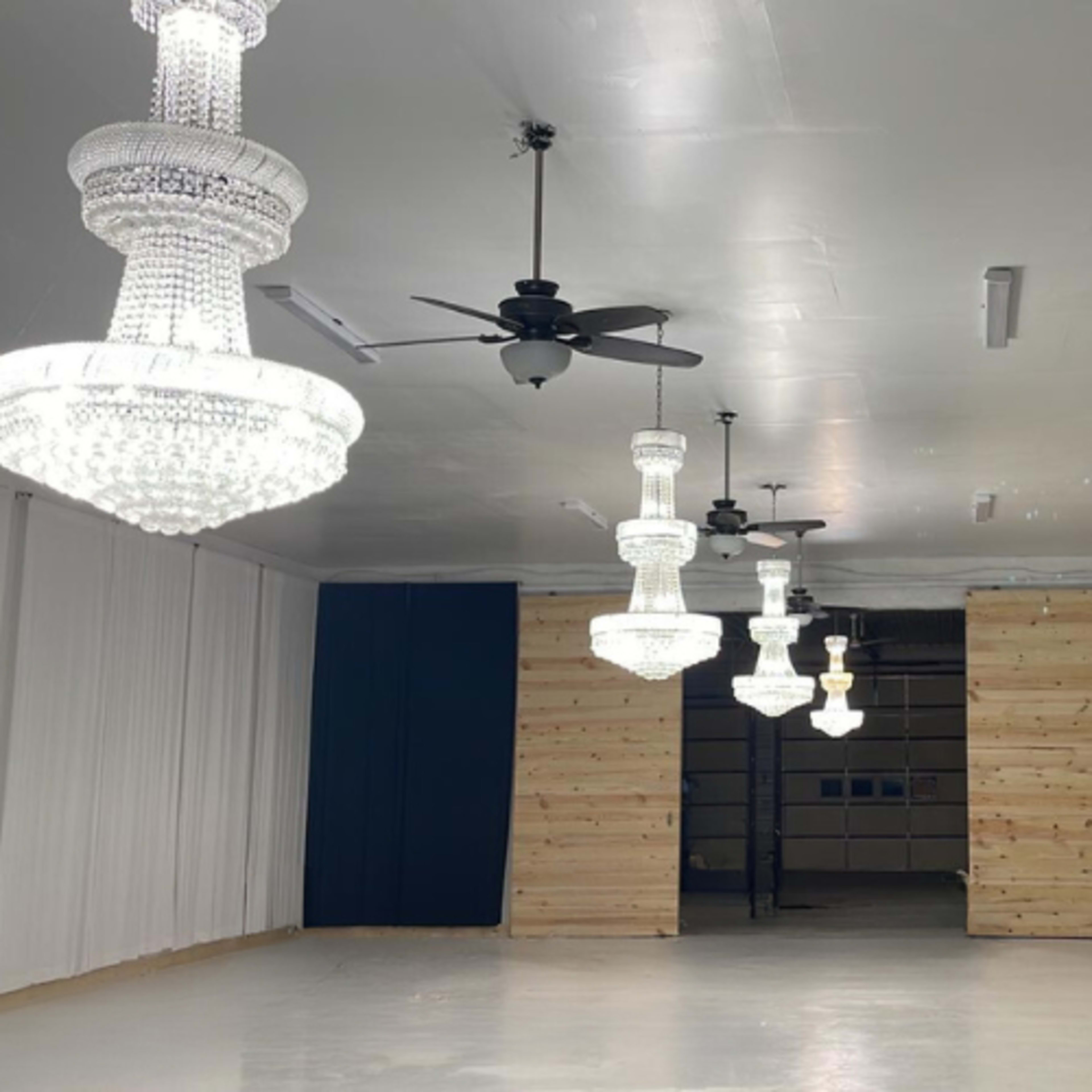 The image shows a well-lit interior space featuring three large chandeliers and ceiling fans, with wooden paneling on one wall.