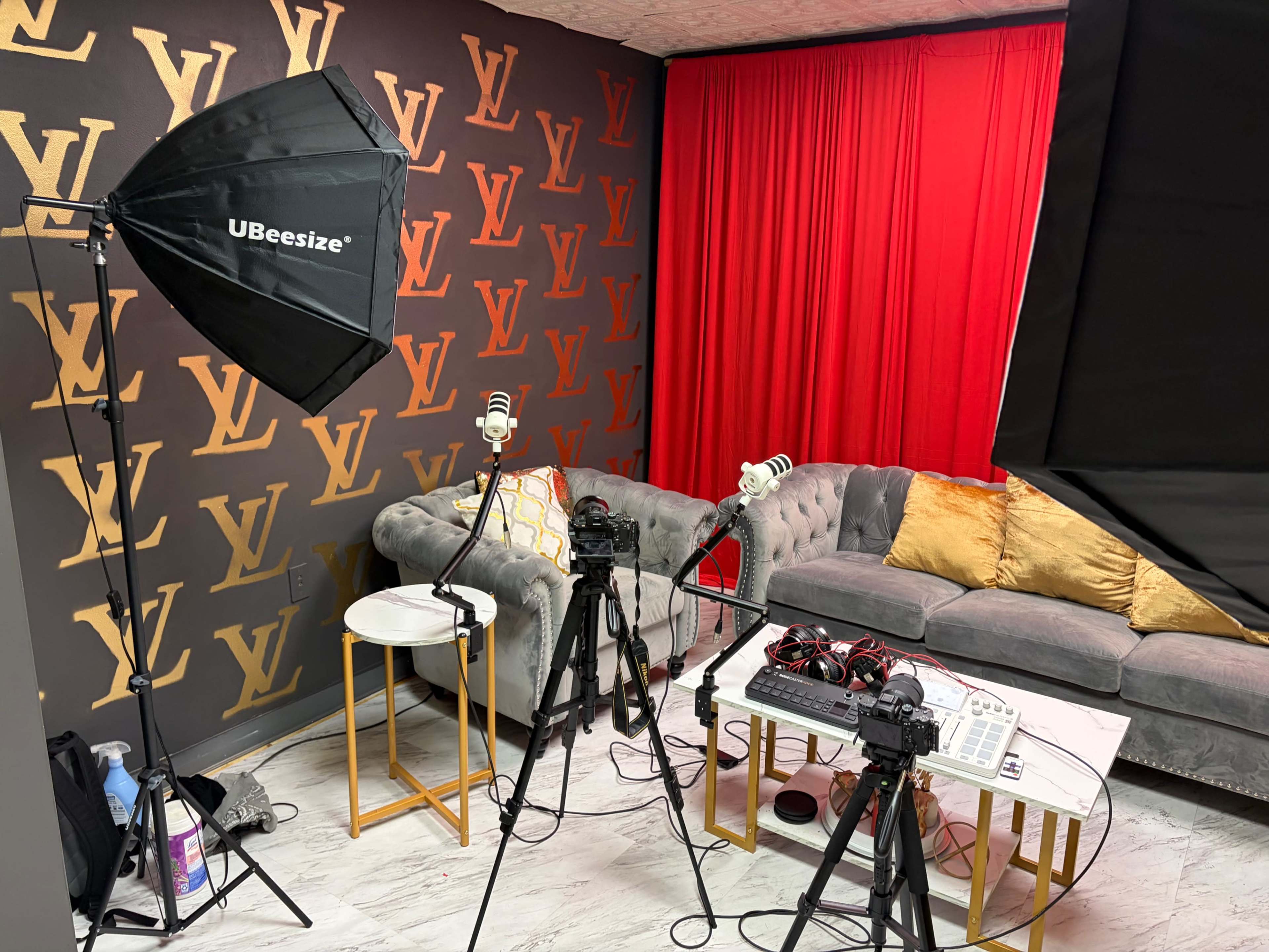 The image shows a recording studio setup with a gray couch, red curtains, and two tripods with microphones and audio equipment.