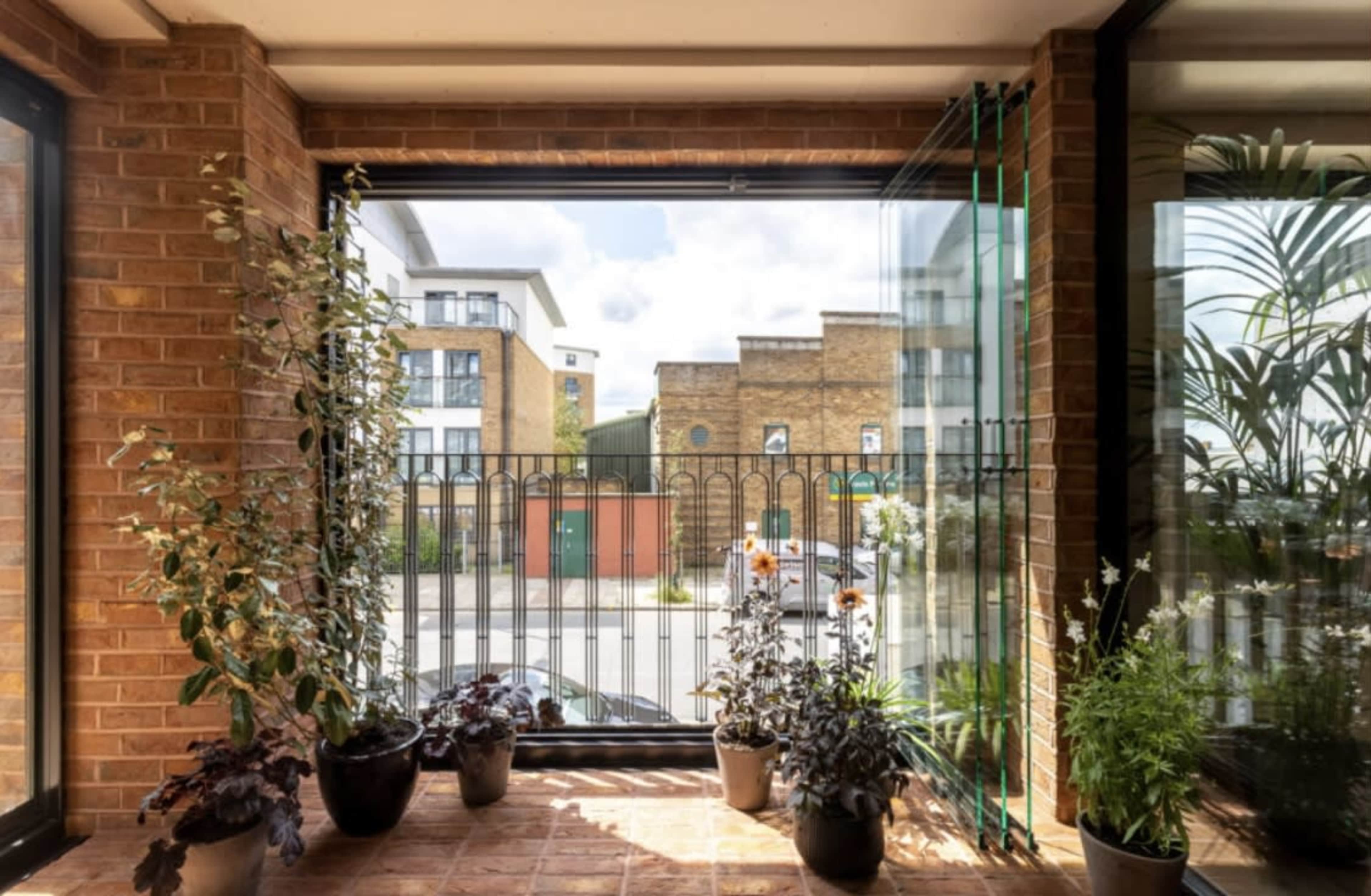 A sunlit balcony features potted plants and a view of urban buildings and a fenced street.