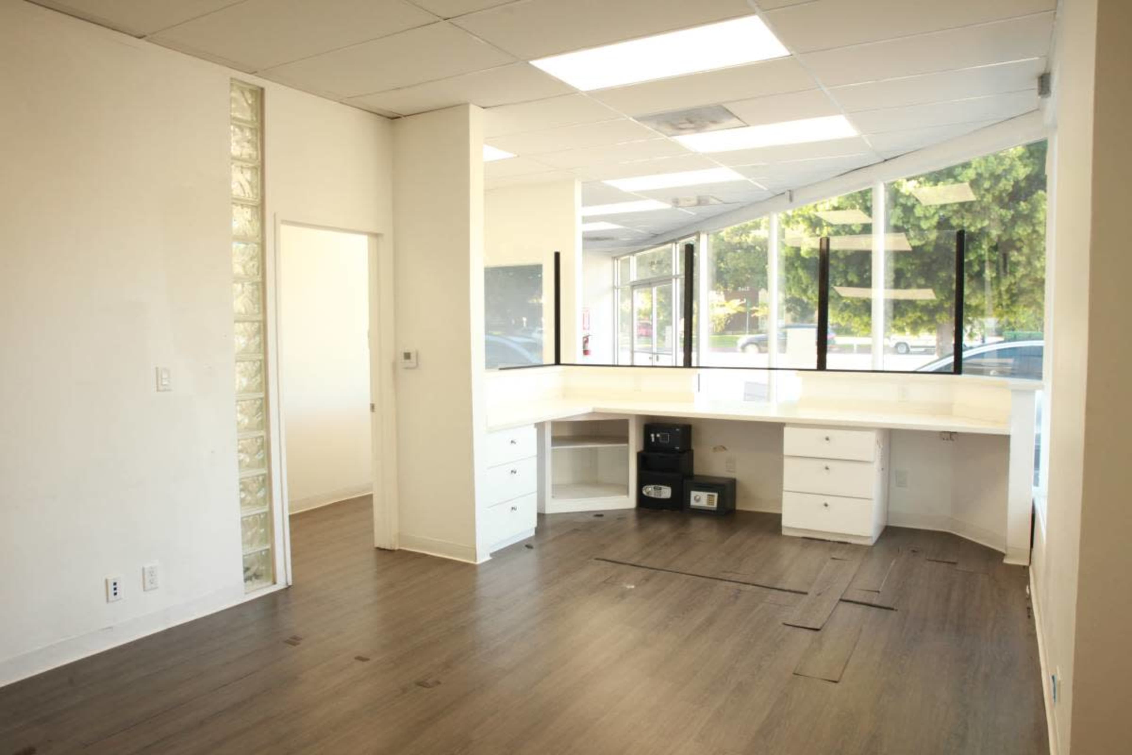 The image shows a vacant office space with a white countertop, built-in drawers, and large windows allowing natural light to enter.