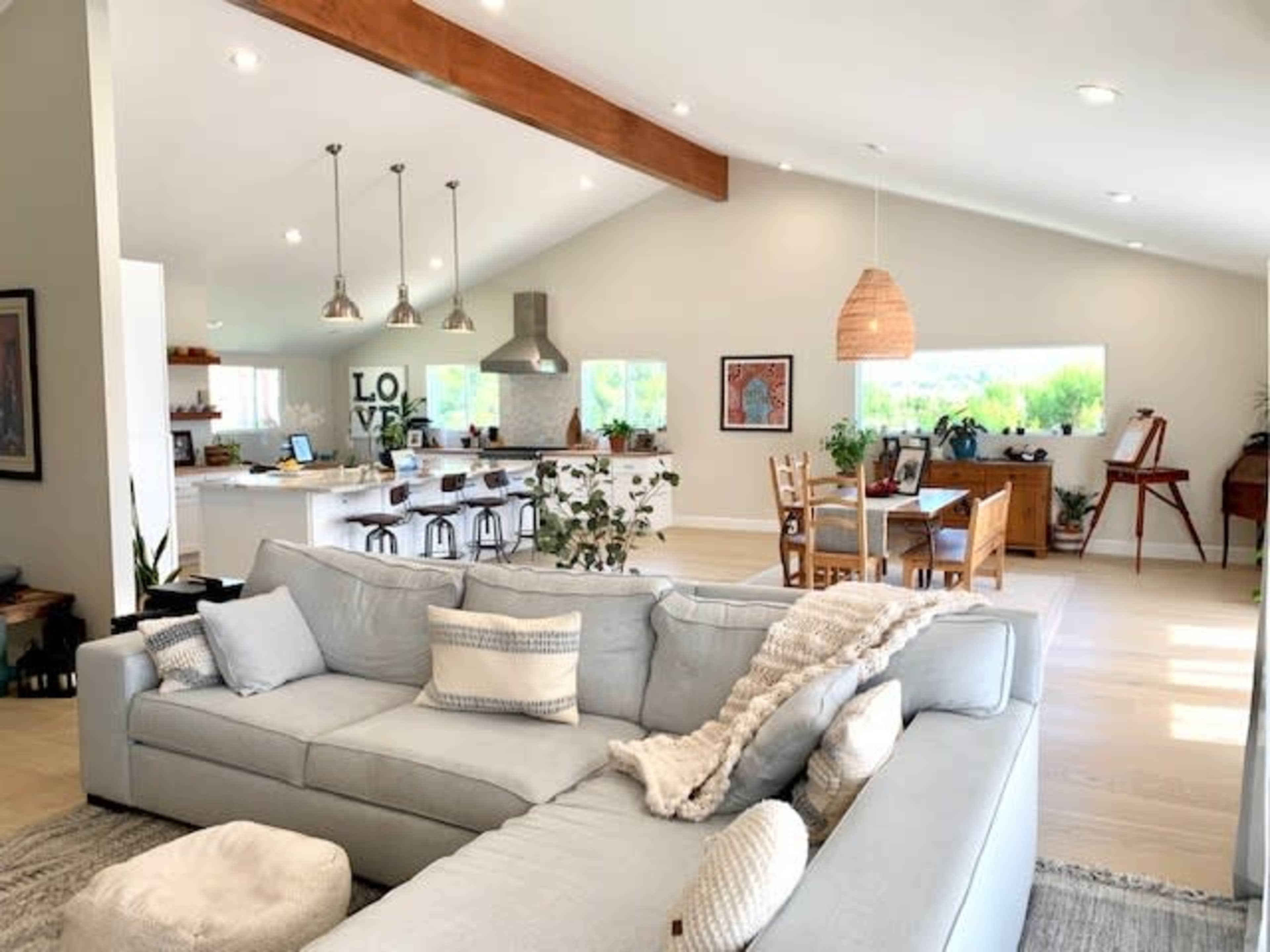 A modern living room with a light-colored sectional sofa, a kitchen in the background with bar seating, and a dining area featuring a wooden table.