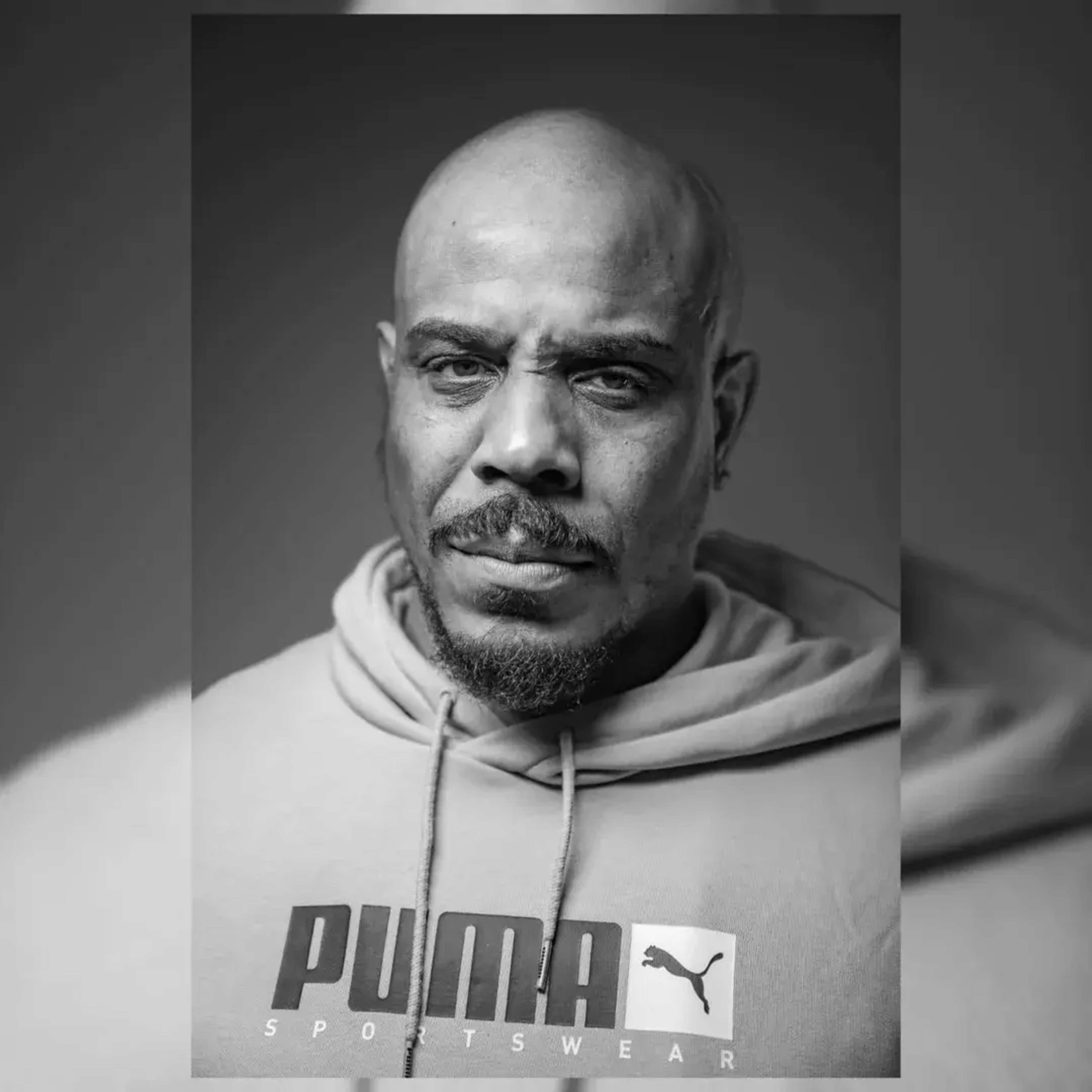 The image shows a man with a shaved head and a serious expression, wearing a gray Puma sweatshirt.