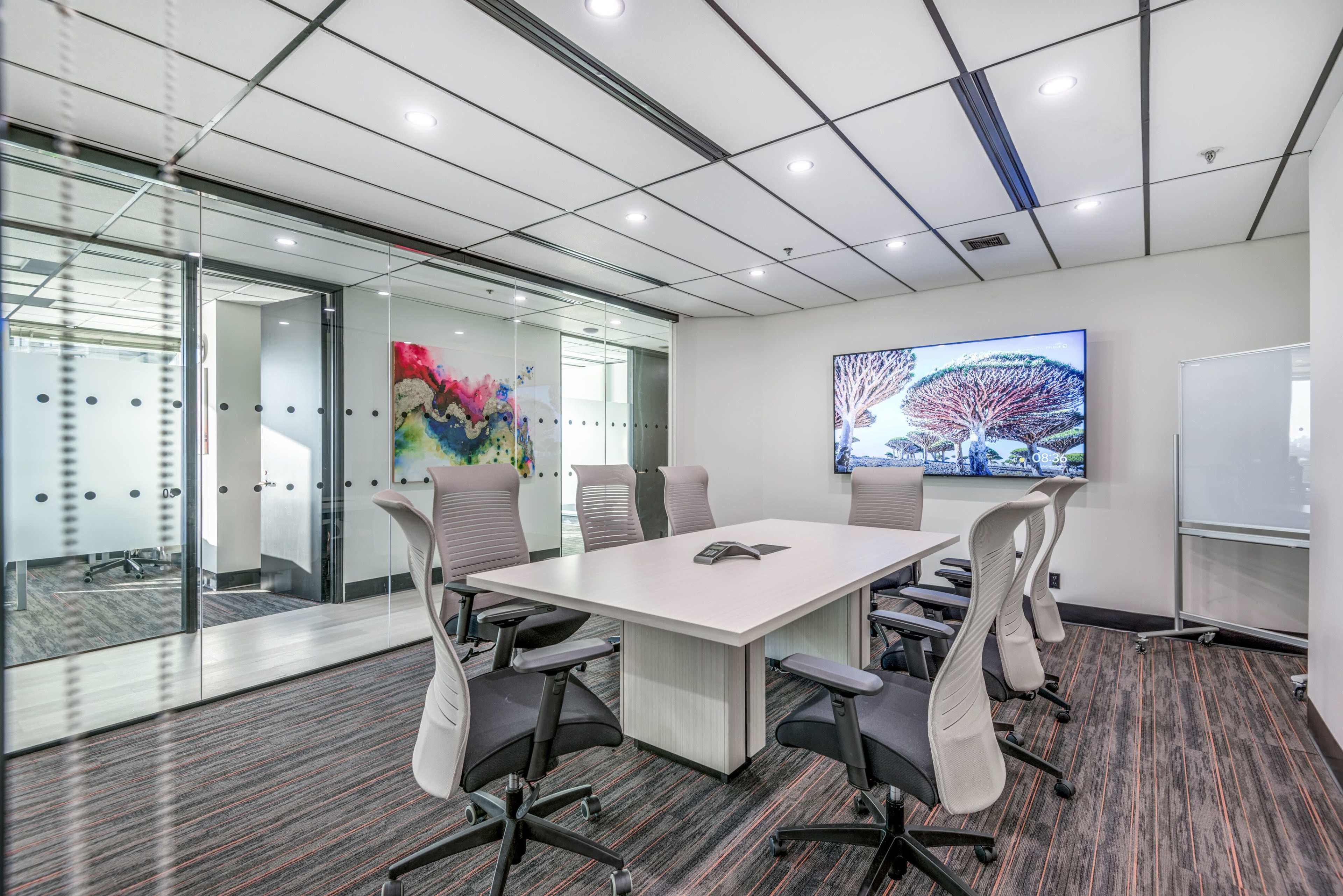 A modern conference room features a wooden table surrounded by six ergonomic chairs, with a large screen displaying a landscape image and glass partitions adorned with colorful artwork.