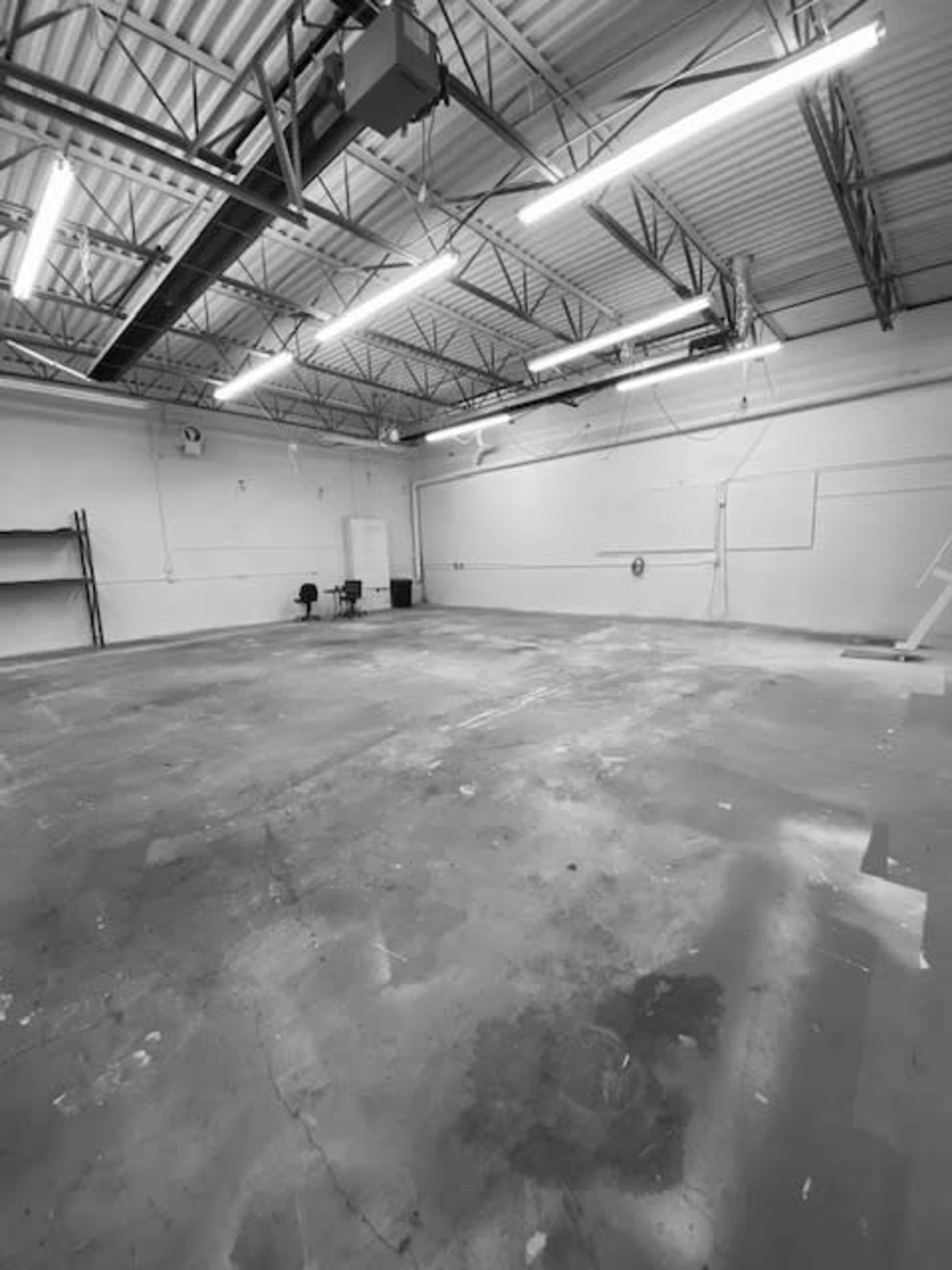 A large, empty room with a concrete floor, overhead lighting, and minimal furnishings in one corner.