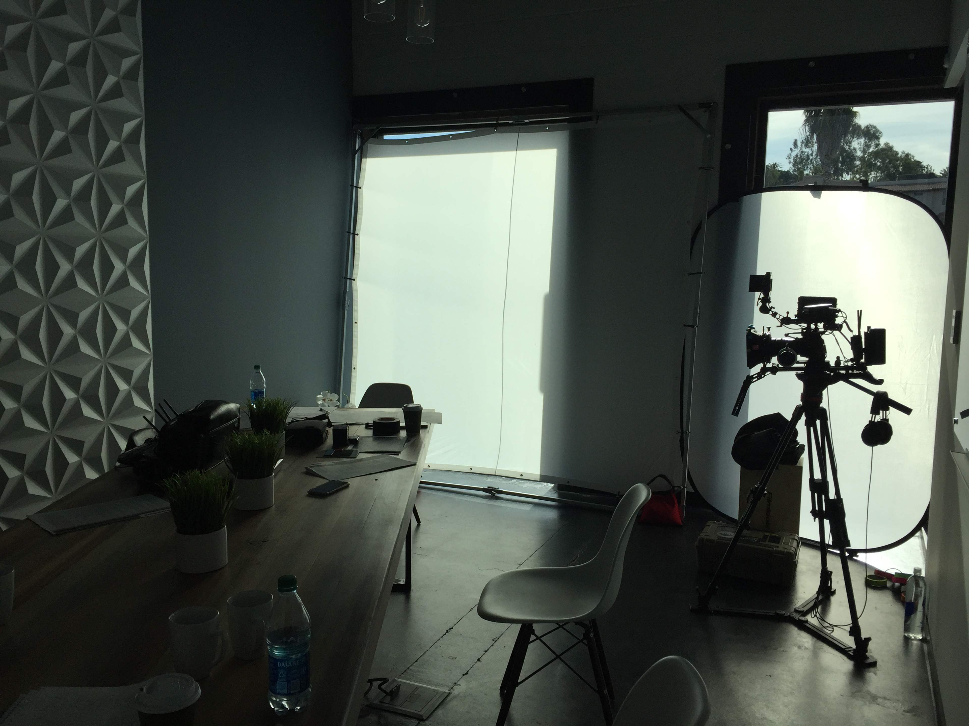 A dimly lit room with a table, chairs, and a camera setup facing a window covered by a white screen.