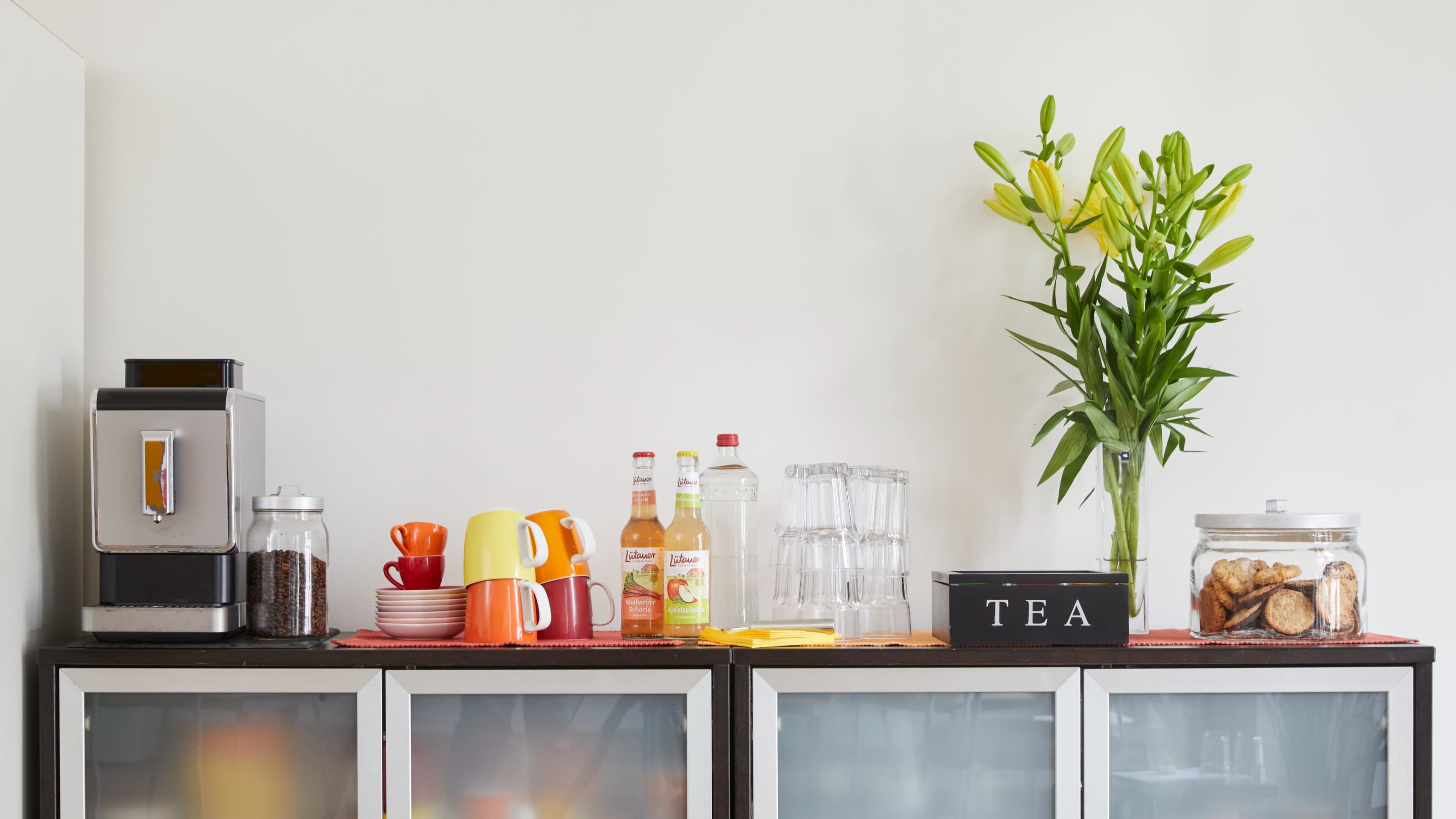 A countertop features a coffee machine, colorful mugs, bottles, glasses, a tea box, and a vase with flowers.