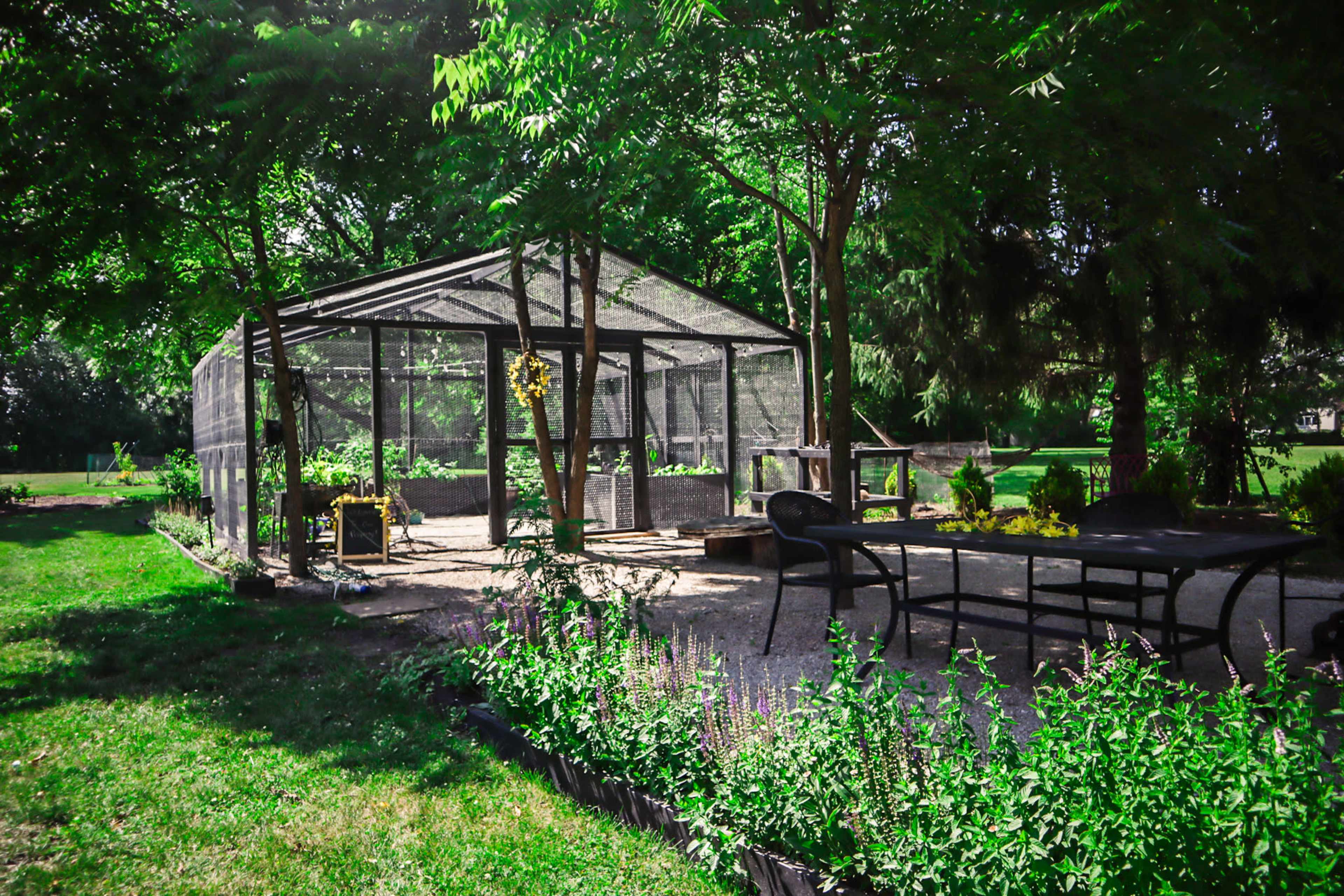 The image shows a garden structure with a wire mesh enclosure surrounded by greenery and a seating area with a table and chairs.