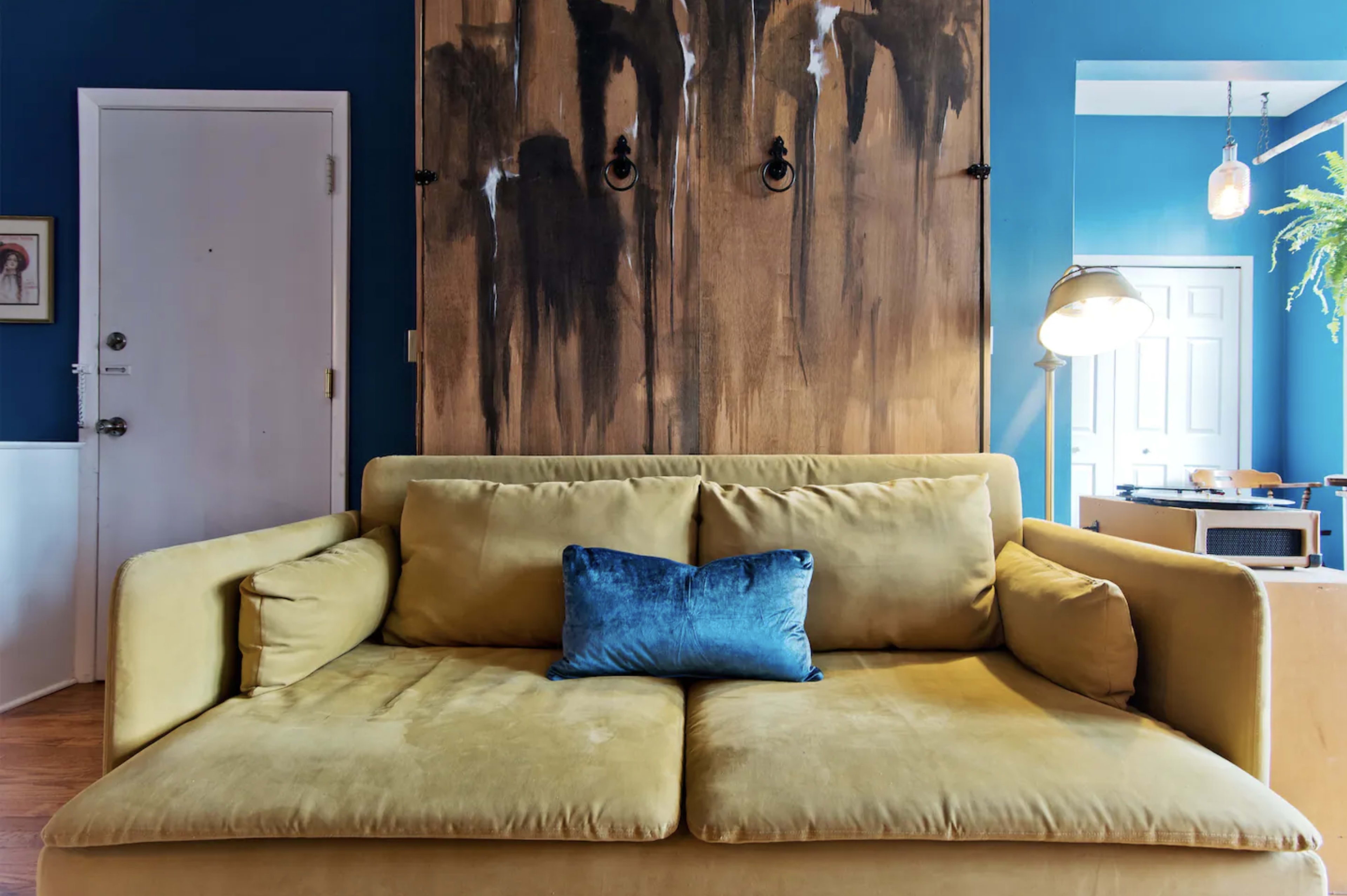 A yellow sofa with a blue cushion is positioned against a blue wall featuring a large, abstract artwork.