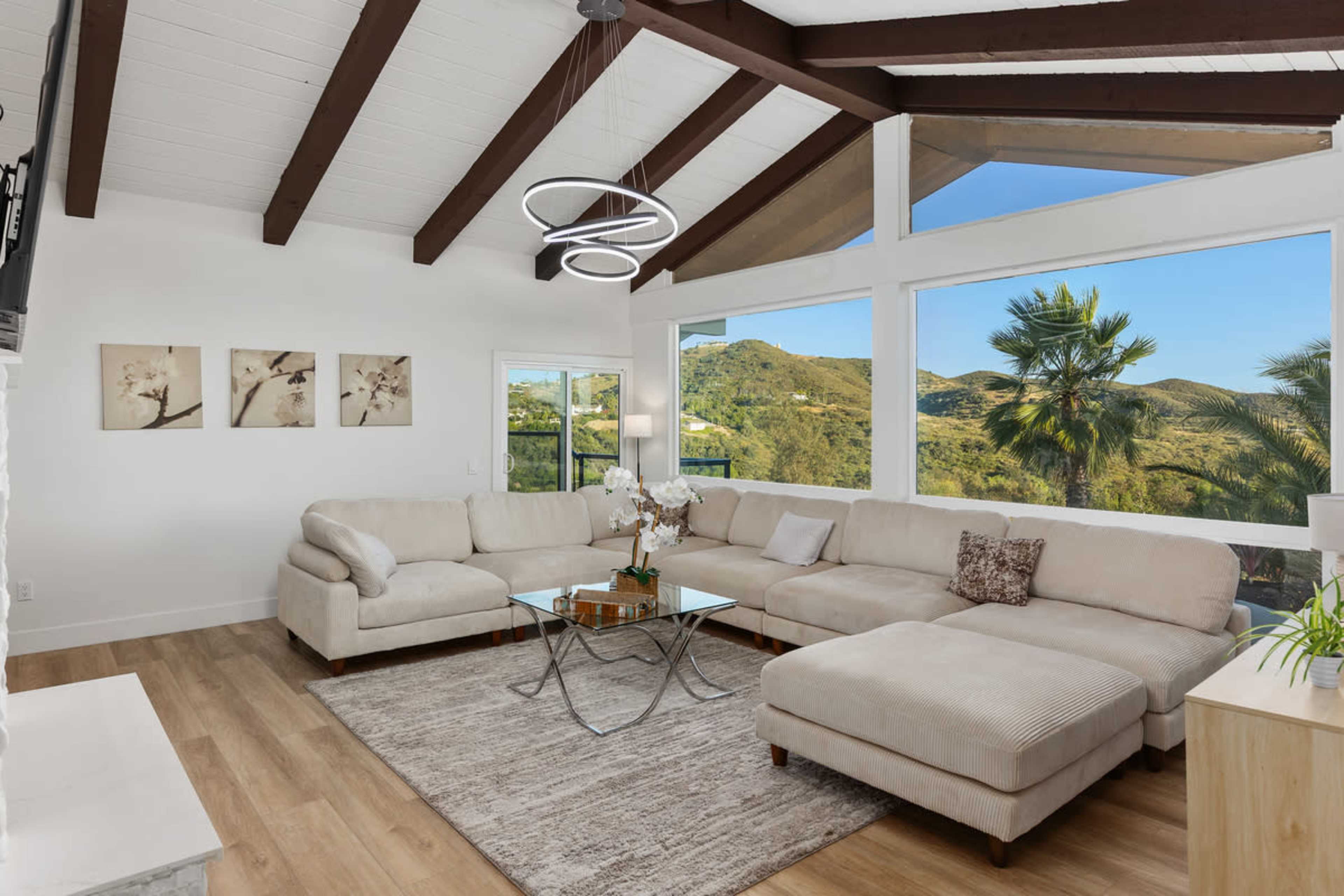 The image shows a modern living room with a large sectional sofa, a glass coffee table, and large windows offering a view of a hilly outdoor landscape.