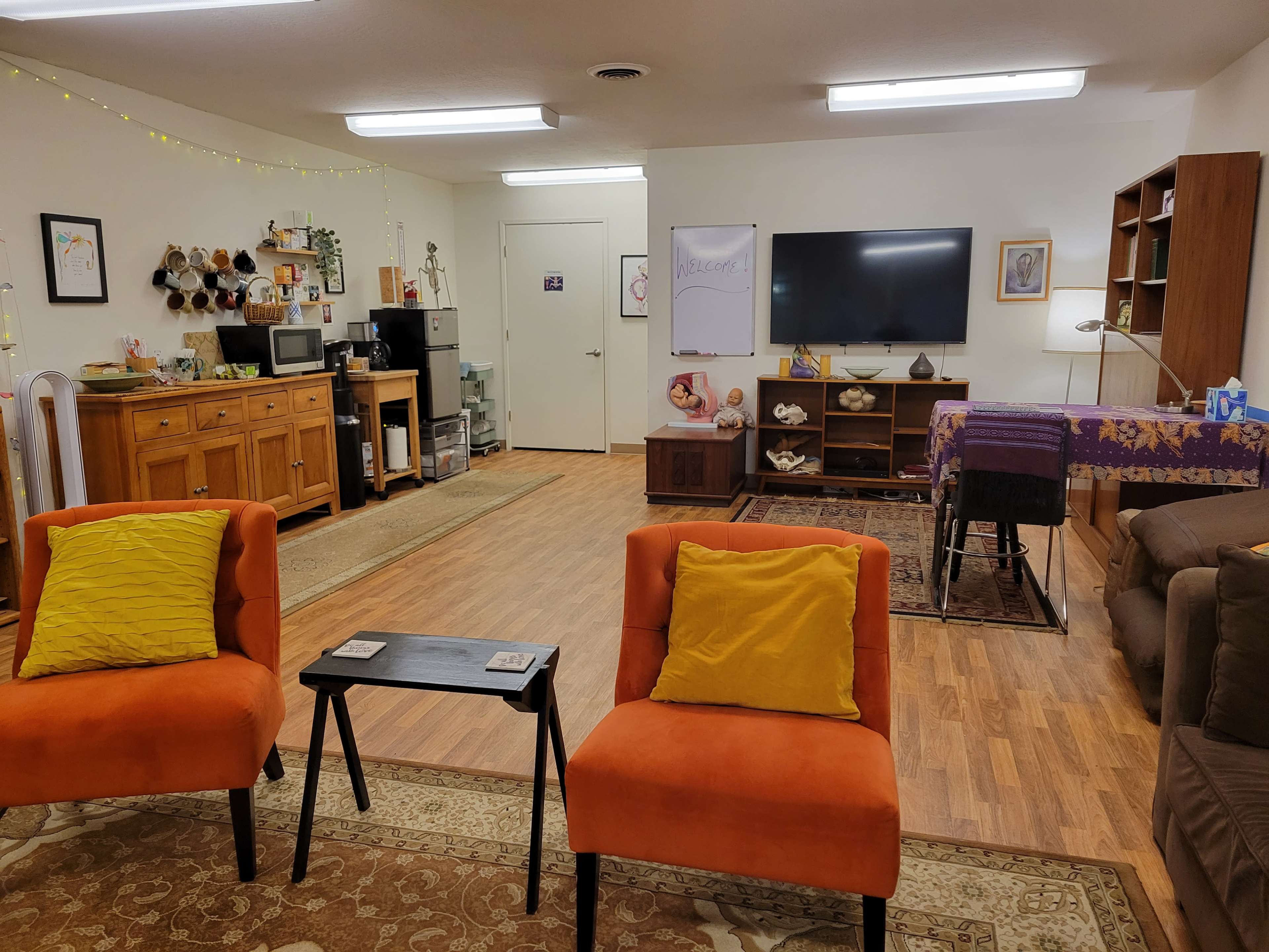 A spacious room with a kitchenette, seating area featuring orange chairs, a dining table, and a television on a stand.