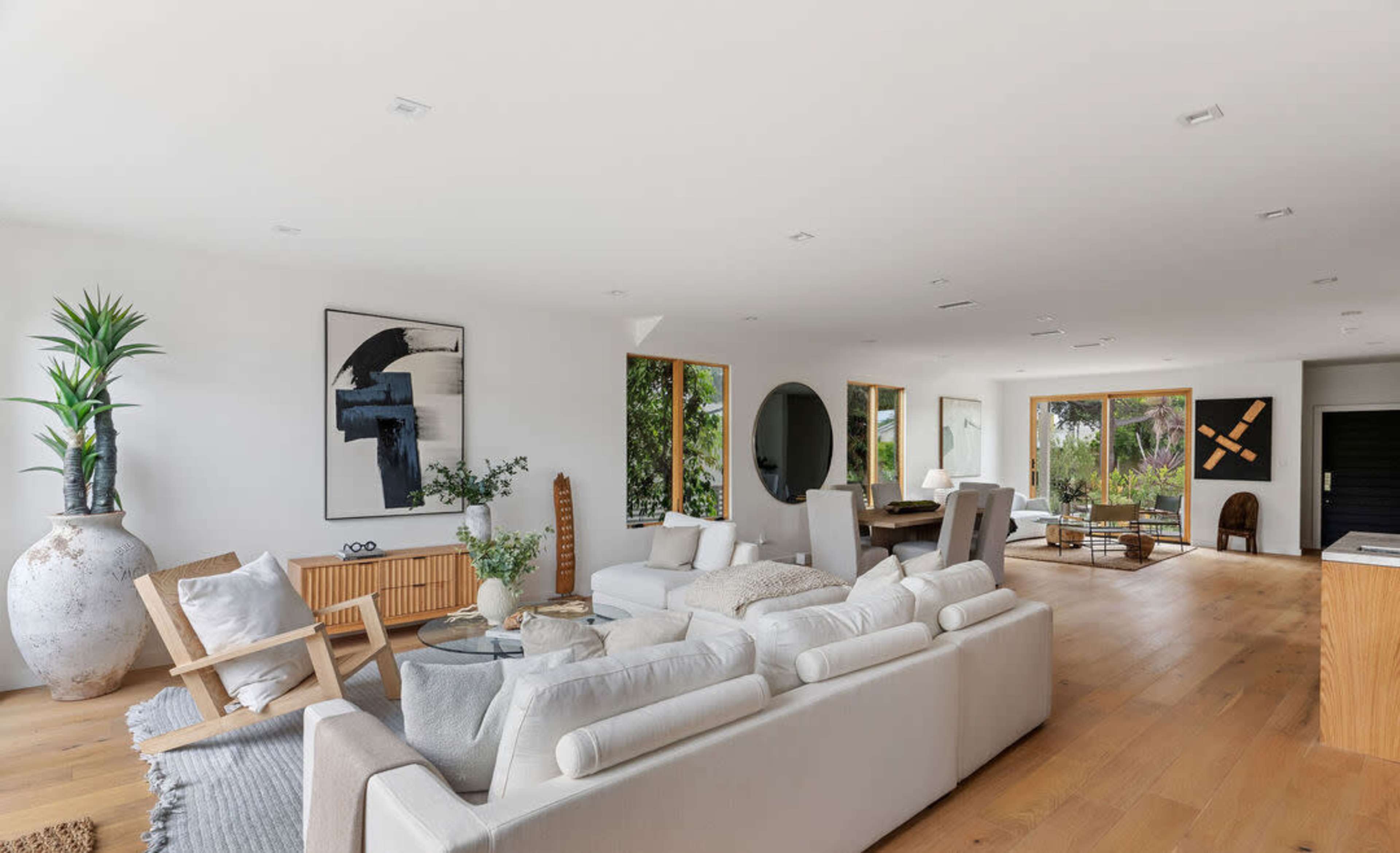 The image showcases a spacious, modern living room featuring a large light-colored sofa, wooden furniture, and various plants, with ample natural light coming through large windows.