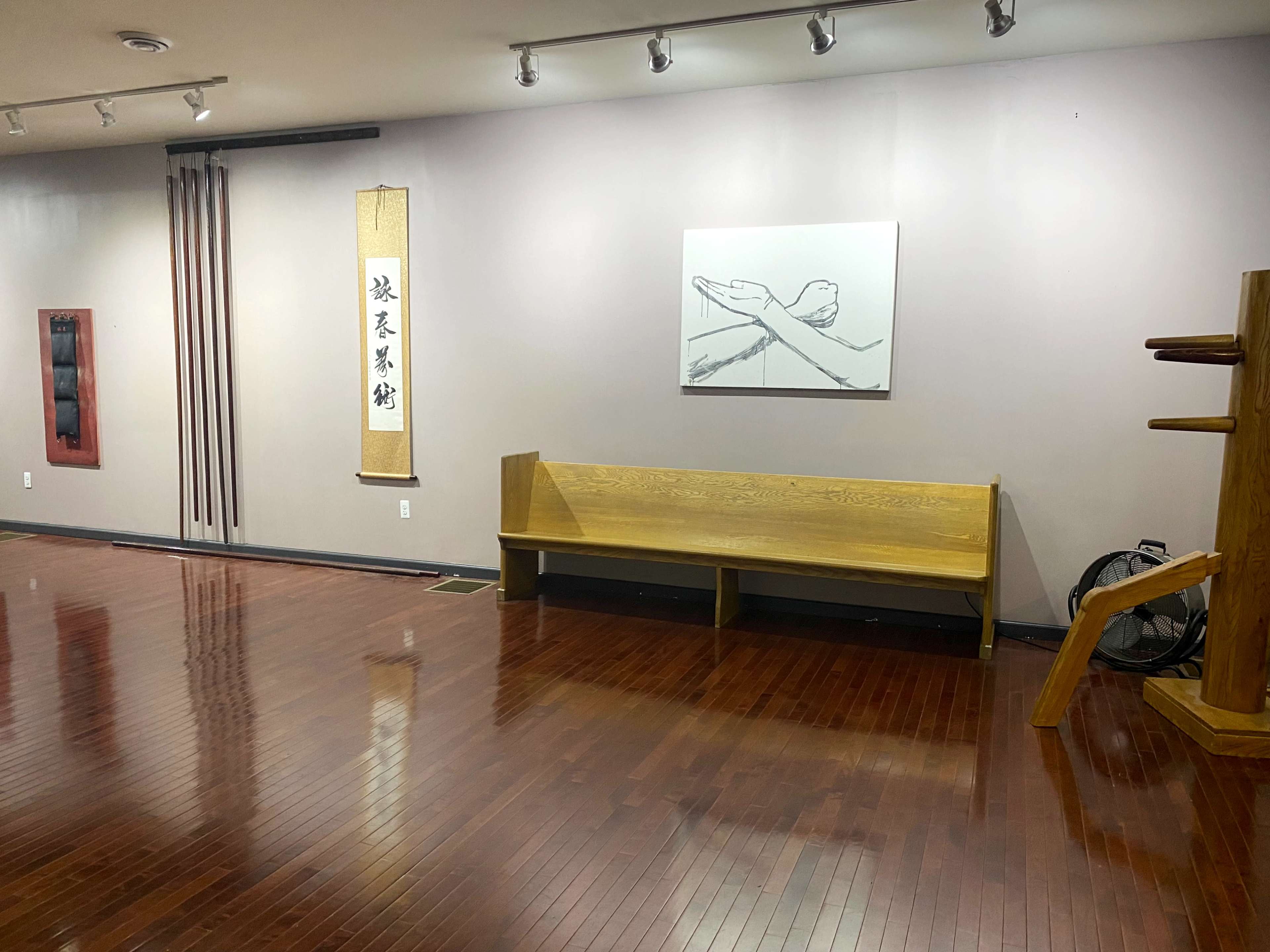 The image shows a spacious room with wooden floors, featuring a bench, martial arts equipment, and a minimalist artwork on the wall.