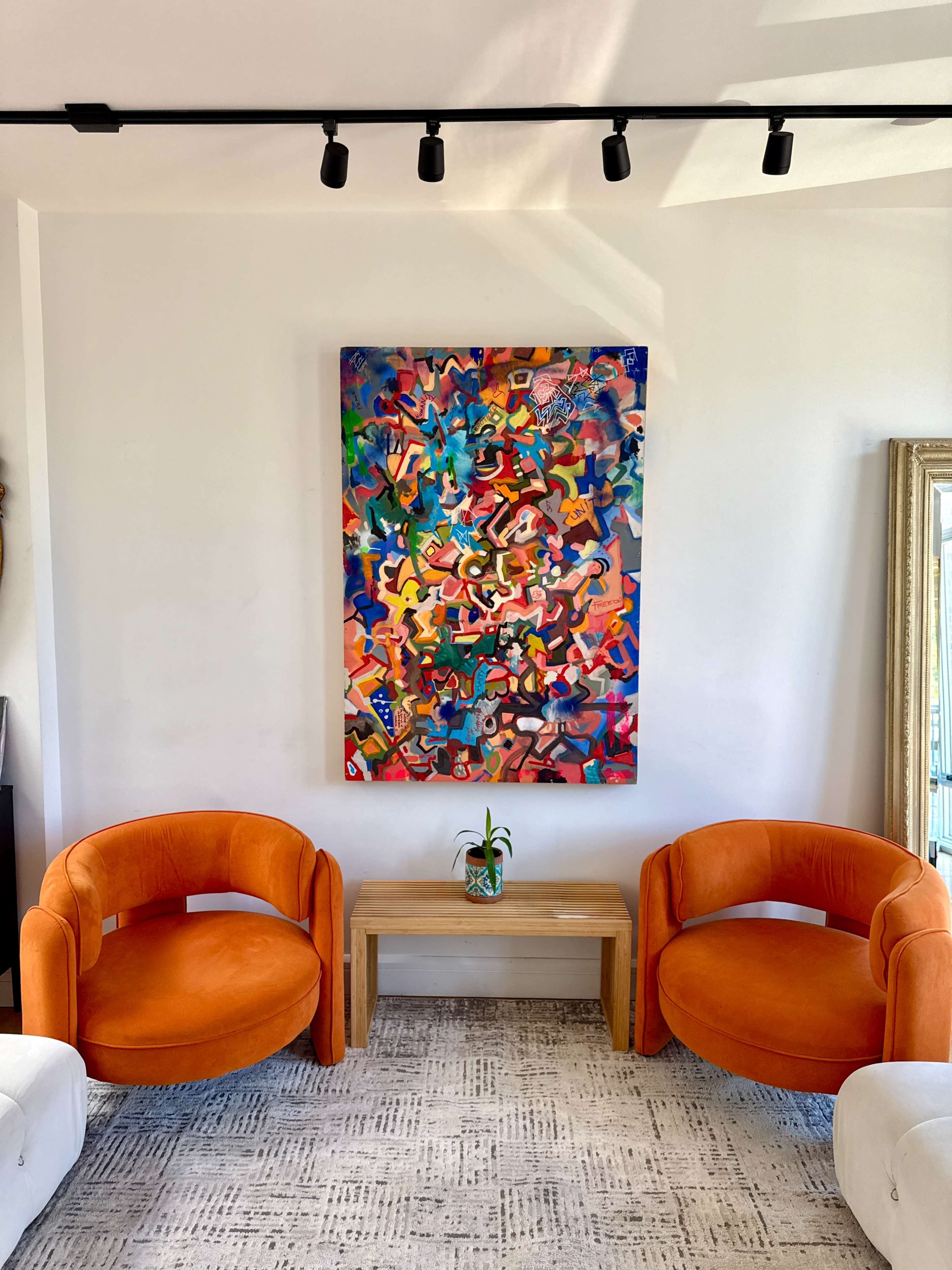 The image features two orange chairs facing each other, a wooden table between them, and a colorful abstract painting mounted on the wall behind.