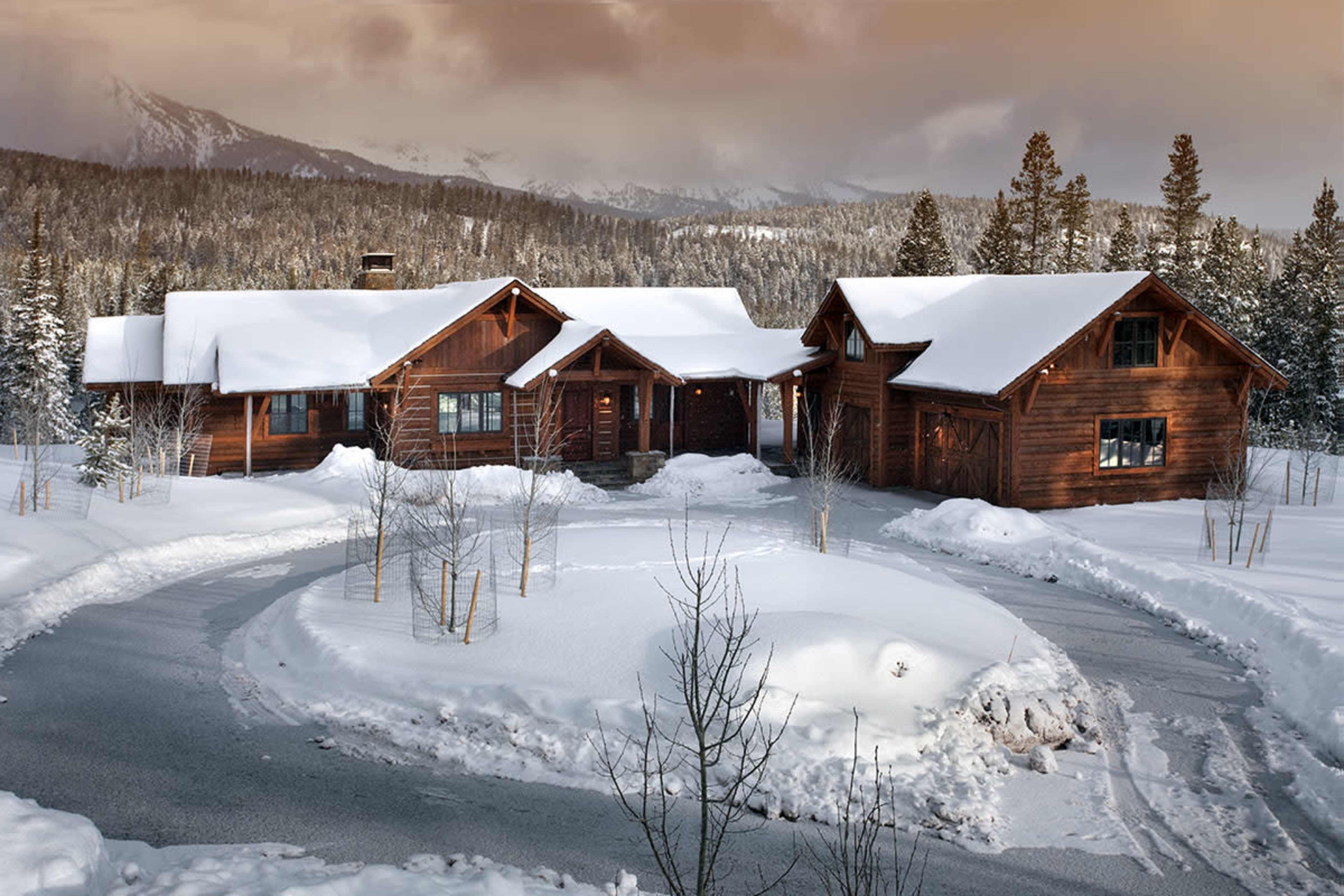 A large wooden house with a sloped roof is surrounded by snow and a winding driveway, set against a backdrop of mountains and evergreen trees.