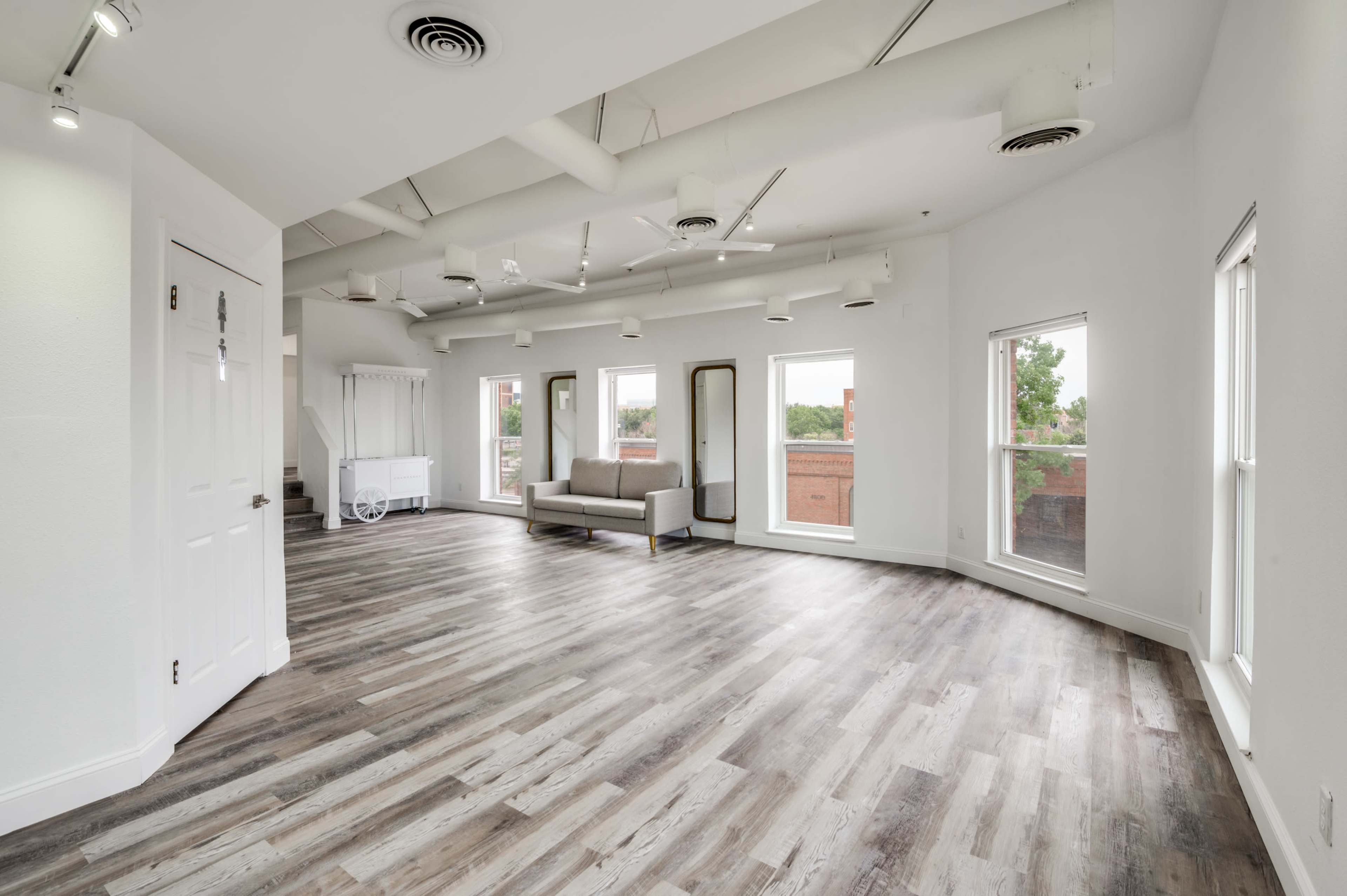 The image shows a spacious, empty room with a light wood floor, large windows, and minimal furnishings including a sofa and a mirror.