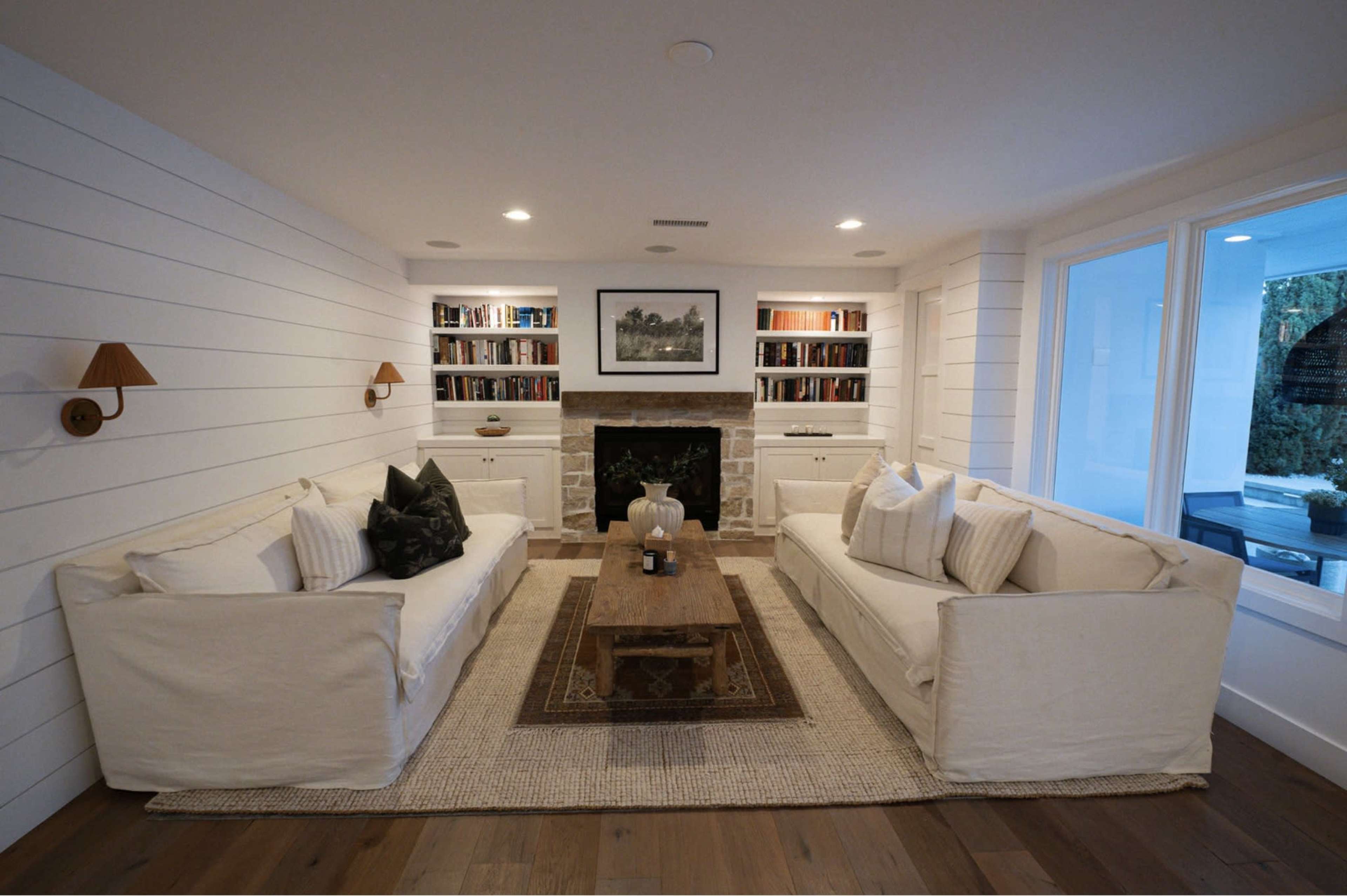 The image shows a modern living room with two white sofas facing a coffee table, a stone fireplace, and built-in bookshelves.