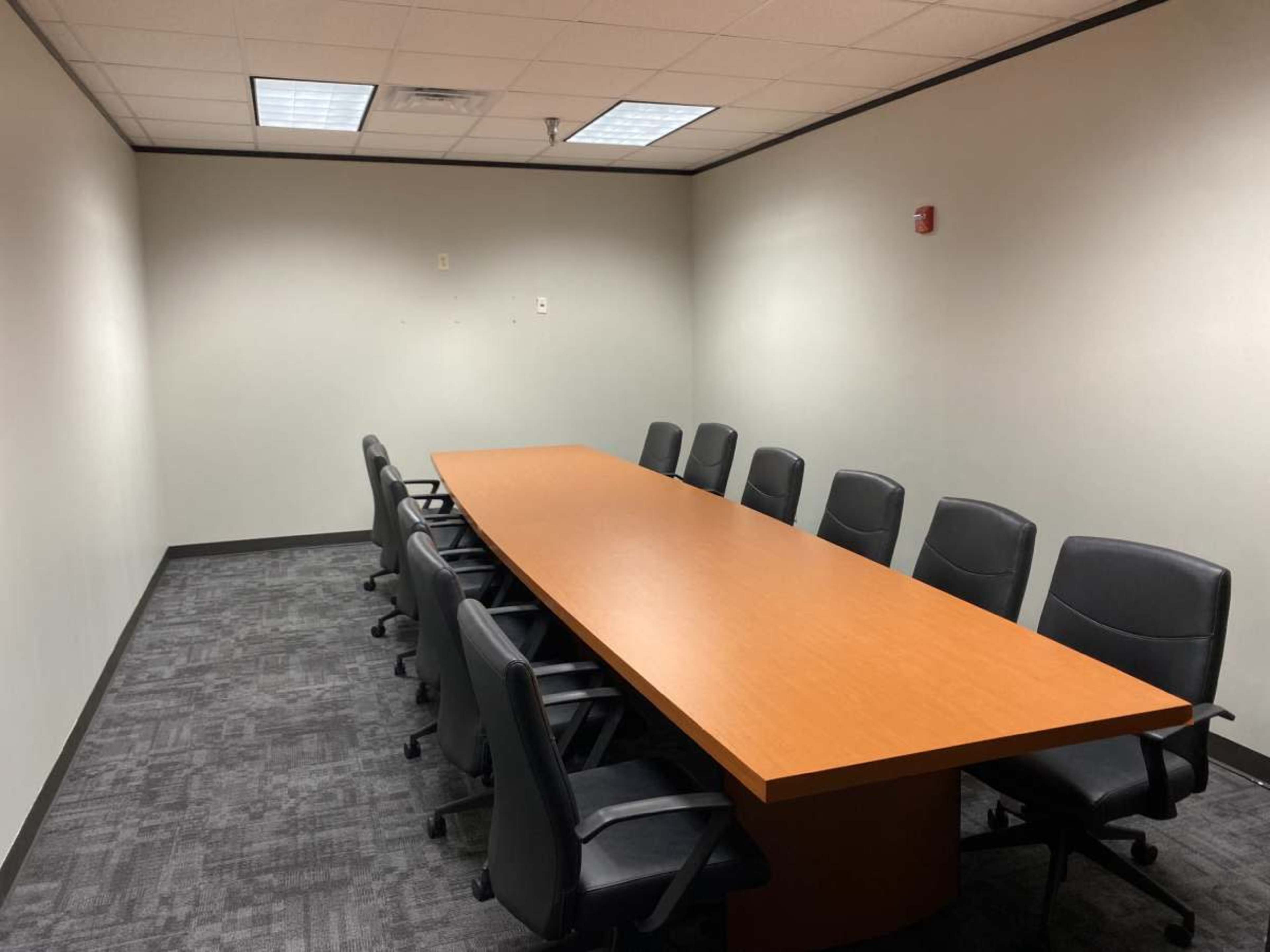 The image shows a conference room with a long wooden table and several black chairs arranged around it, set against plain walls and carpeted flooring.