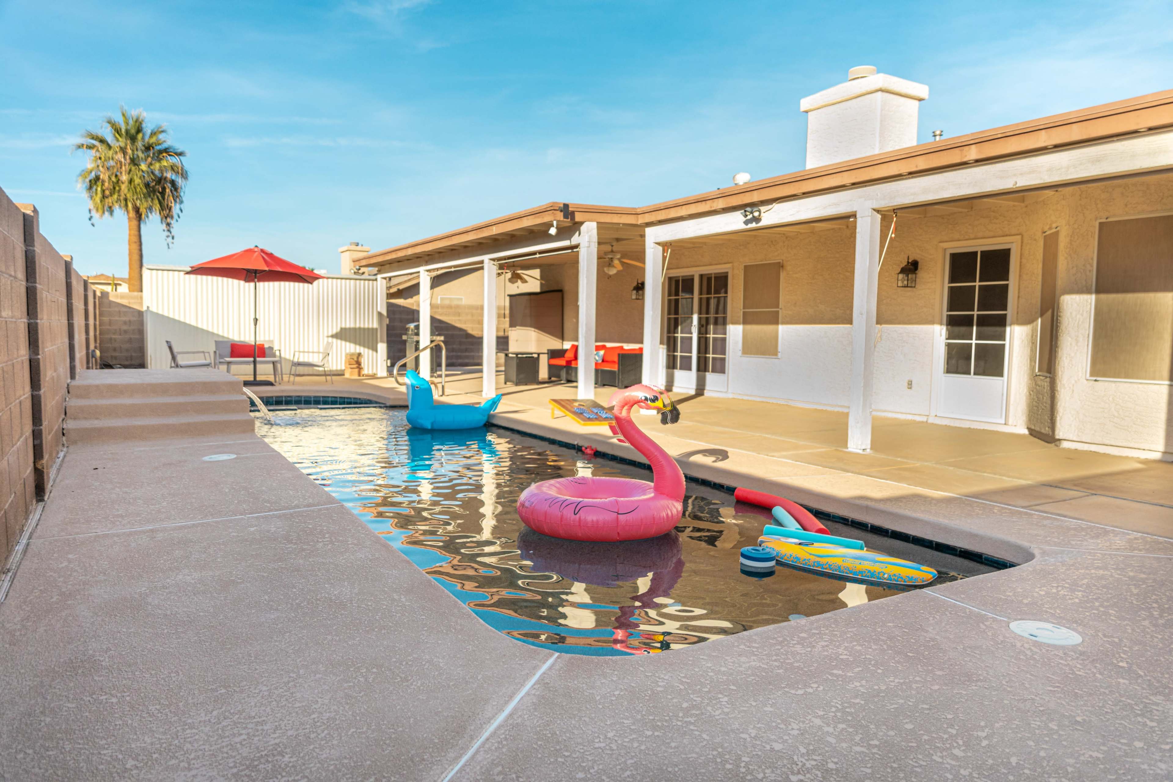 A swimming pool with colorful floats, surrounded by a patio and a house, under a clear blue sky.